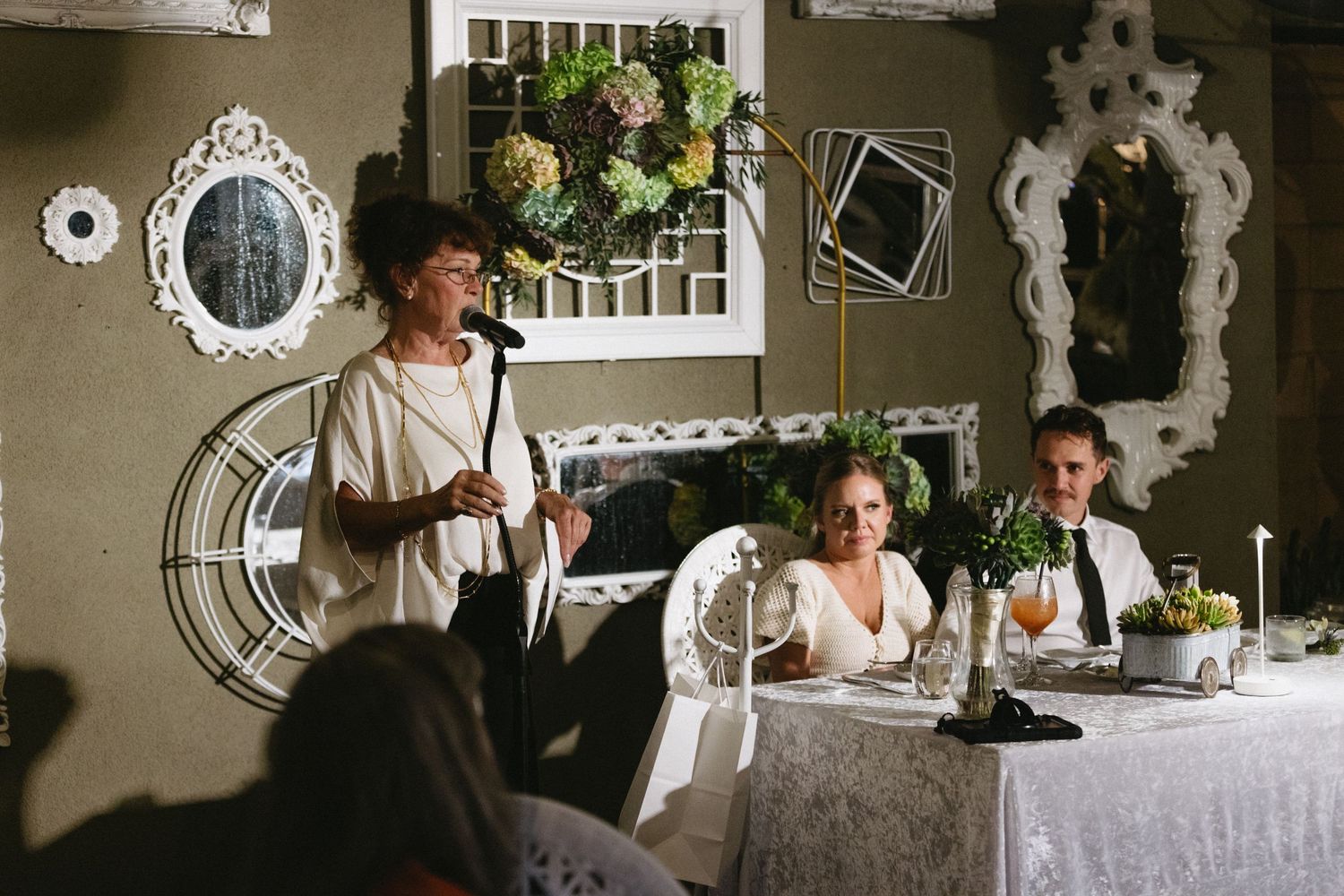 Woman giving a speech at an elegant wedding reception with decorative mirrors and floral arrangements.