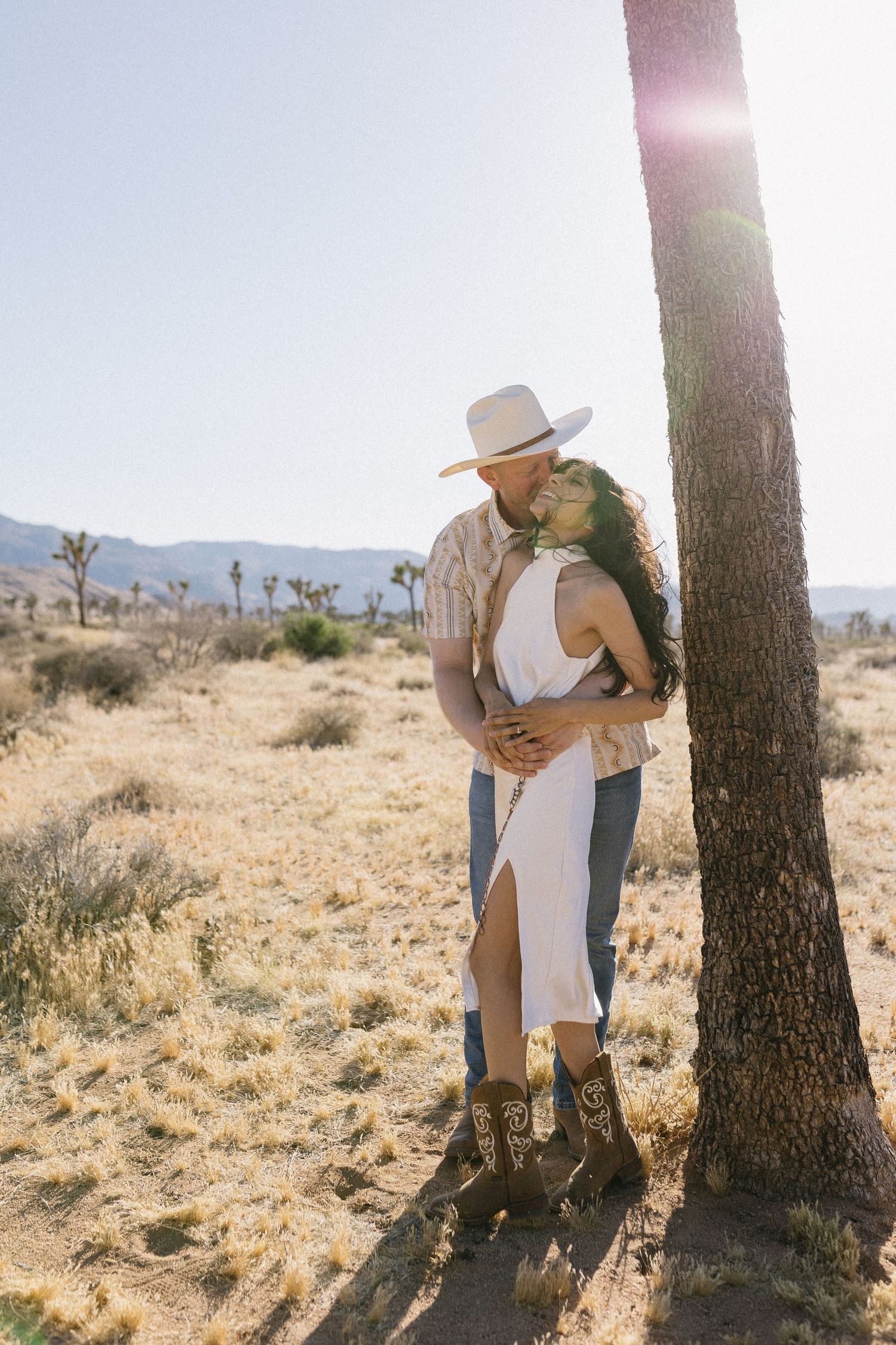 Couple embracing against a tree in desert landscape with Joshua trees in background.