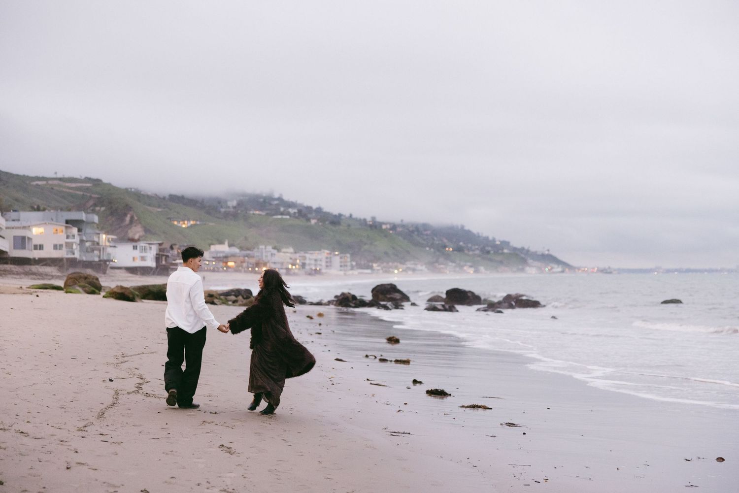 Couple walking hand in hand along misty beach with rocks.