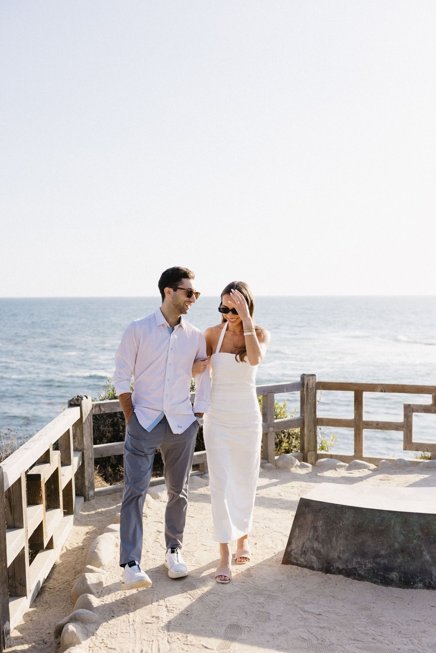 Couple in white outfits walking along a coastal boardwalk overlooking the ocean.
