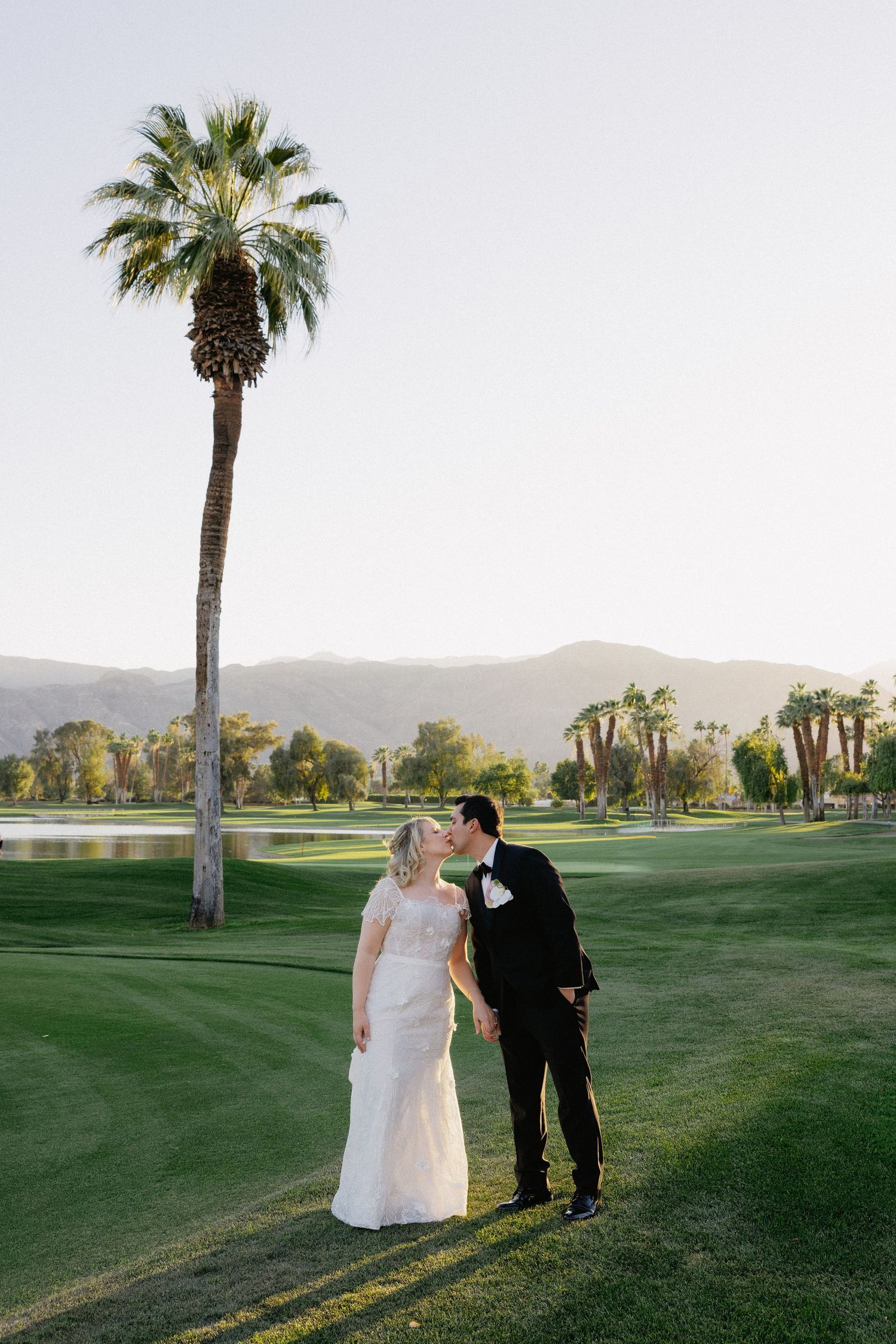 Bride and groom share a romantic kiss on a lush golf course with palm trees and mountains in Palm Springs.