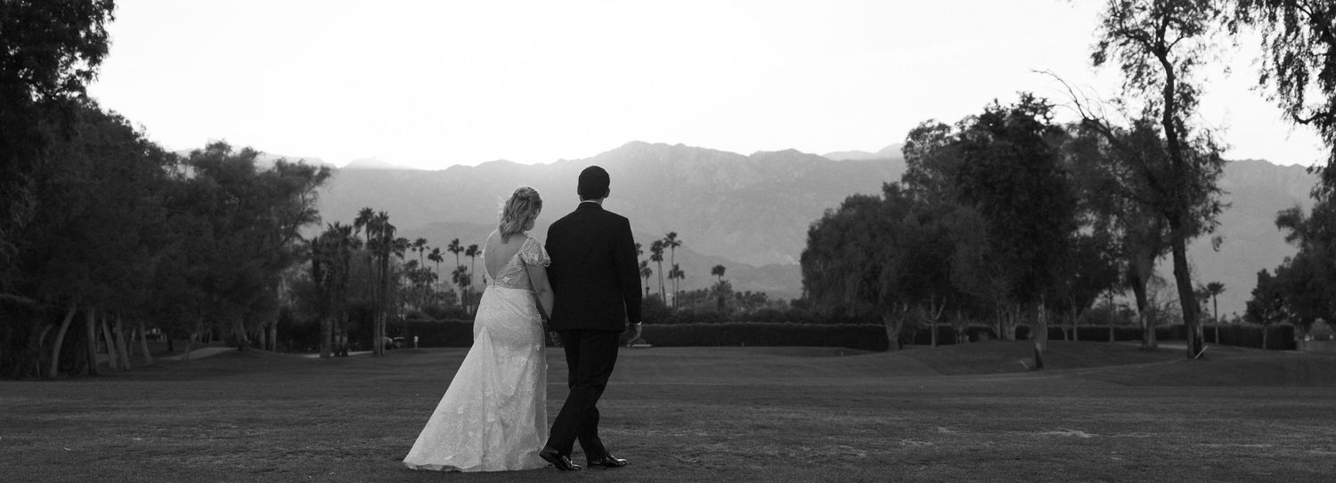 Black and white photo of a bride and groom standing together in a field with mountains in the background.