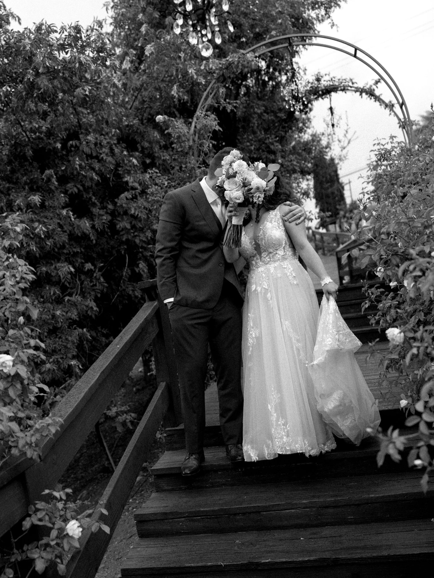 Black and white photo of a bride and groom kissing on wooden steps, surrounded by lush greenery and a floral arch.