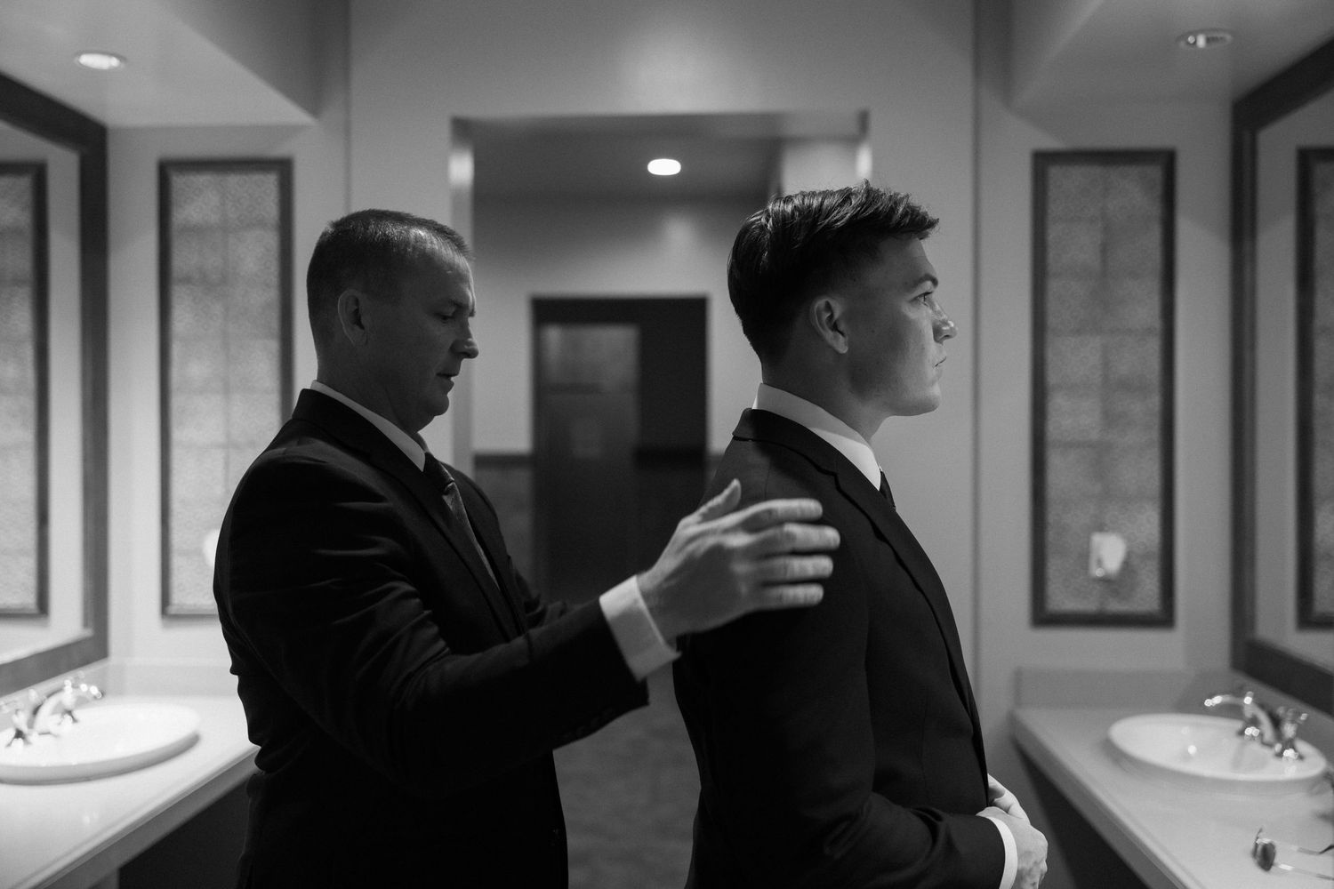 Black and white photo of two men in suits, one adjusting the other's jacket collar in an elegant bathroom.