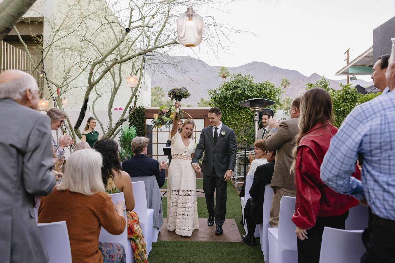 Bride and groom walk down outdoor wedding aisle surrounded by cheering guests with mountain backdrop.