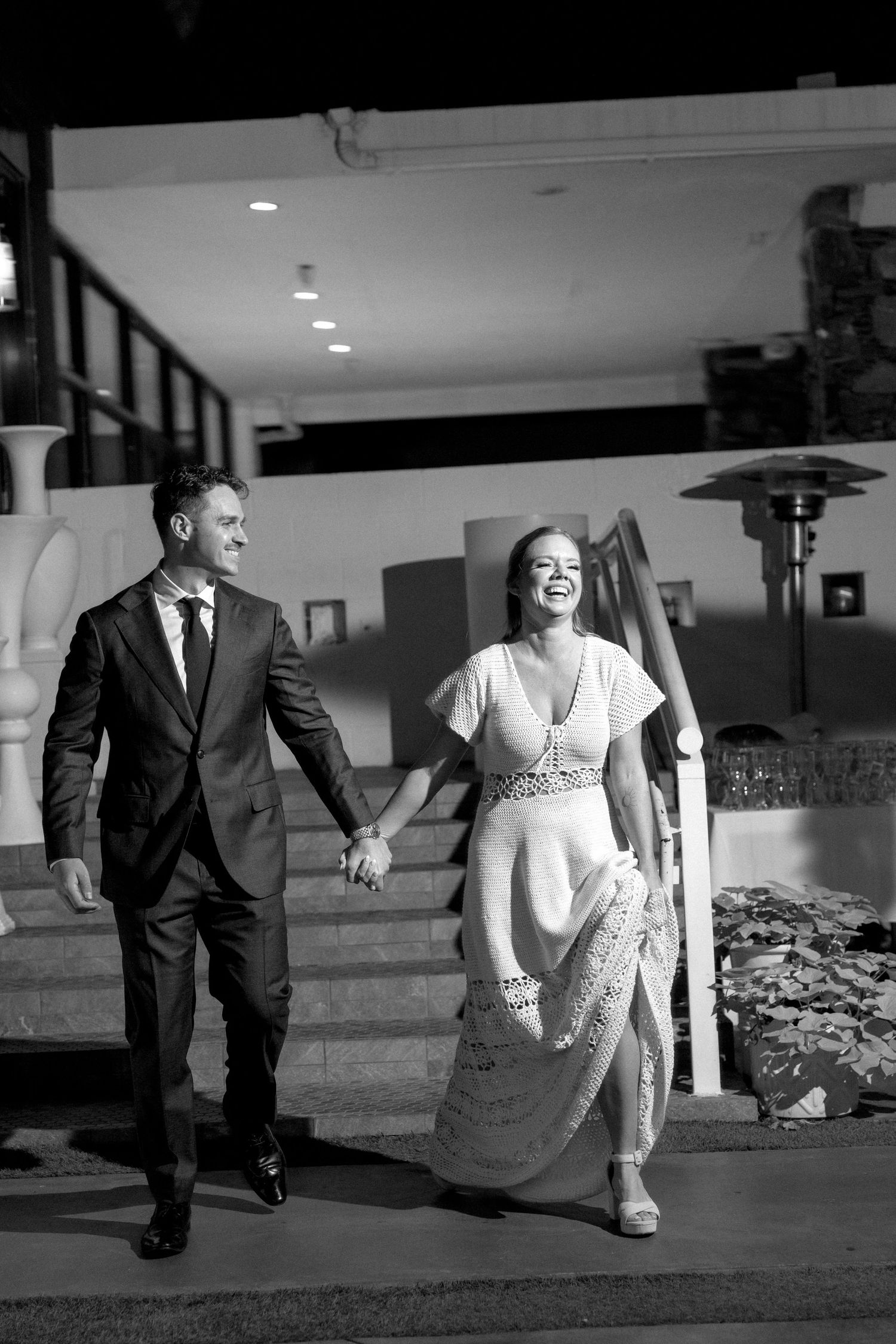 Black and white photo of a smiling couple in formal attire holding hands while walking down outdoor steps at night.