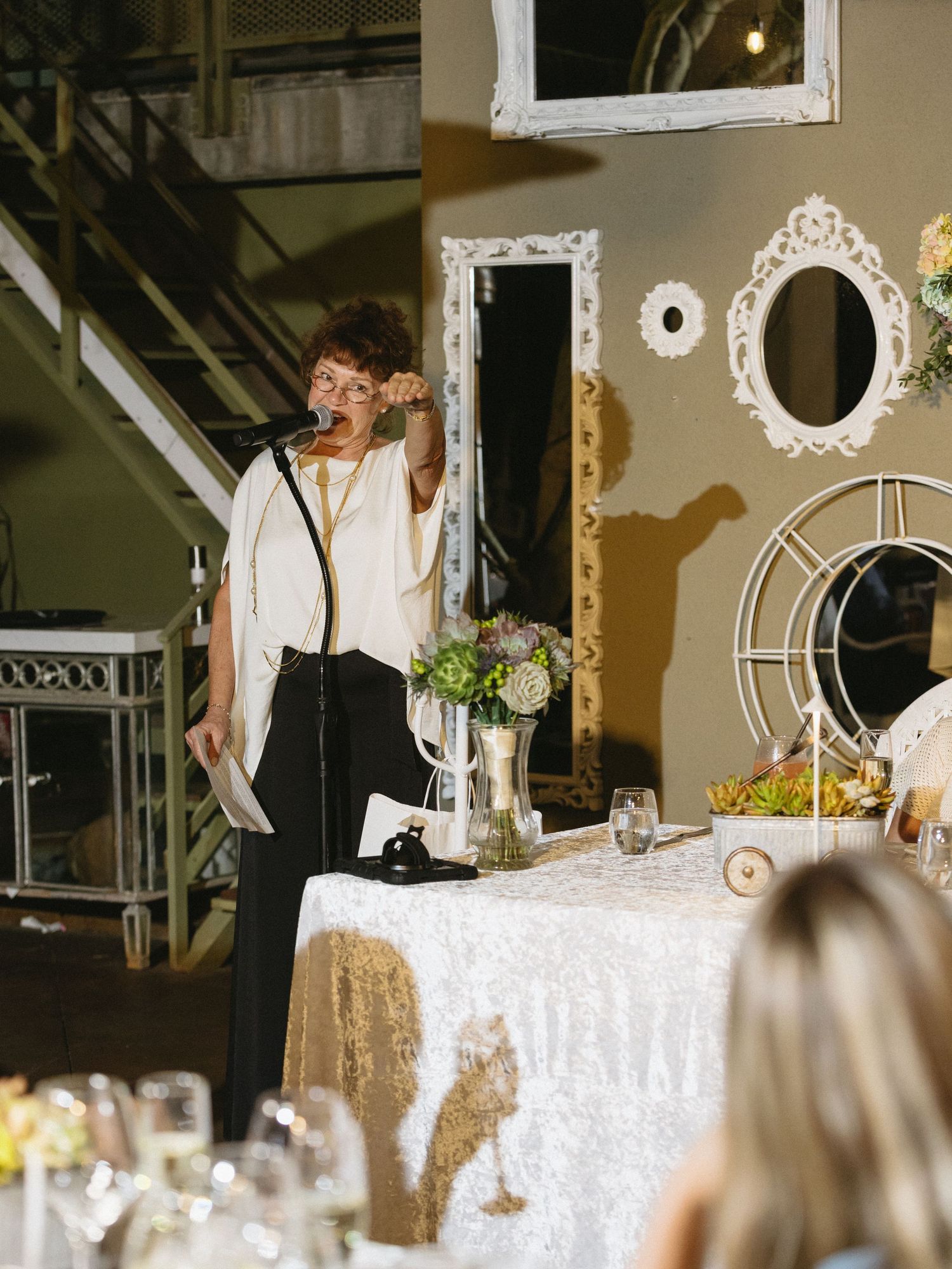 Woman speaking at microphone during elegant indoor event with decorative mirrors and floral centerpieces on sequined tablecloth.