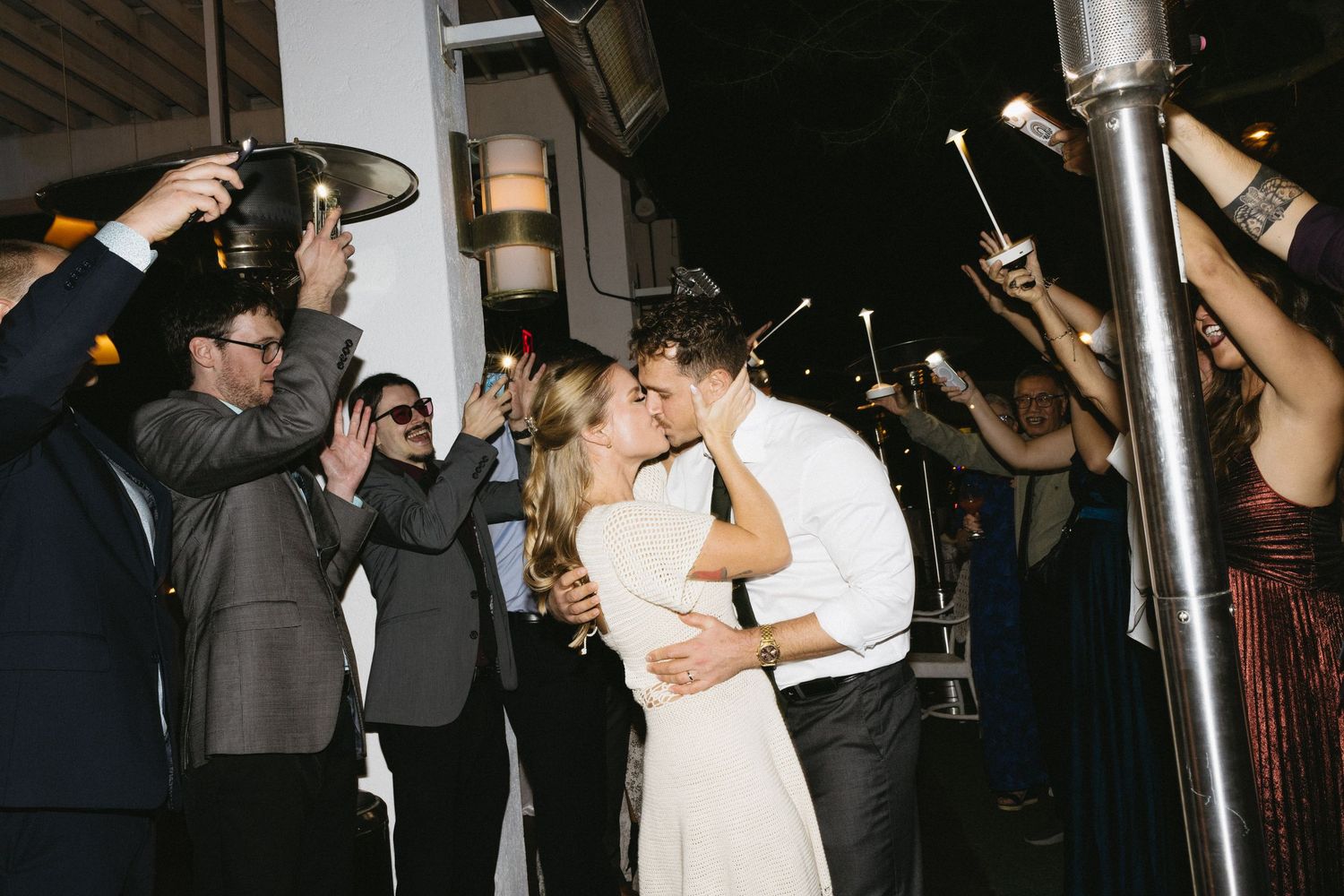 Bride and groom share a kiss surrounded by cheering guests holding sparklers at their wedding reception.