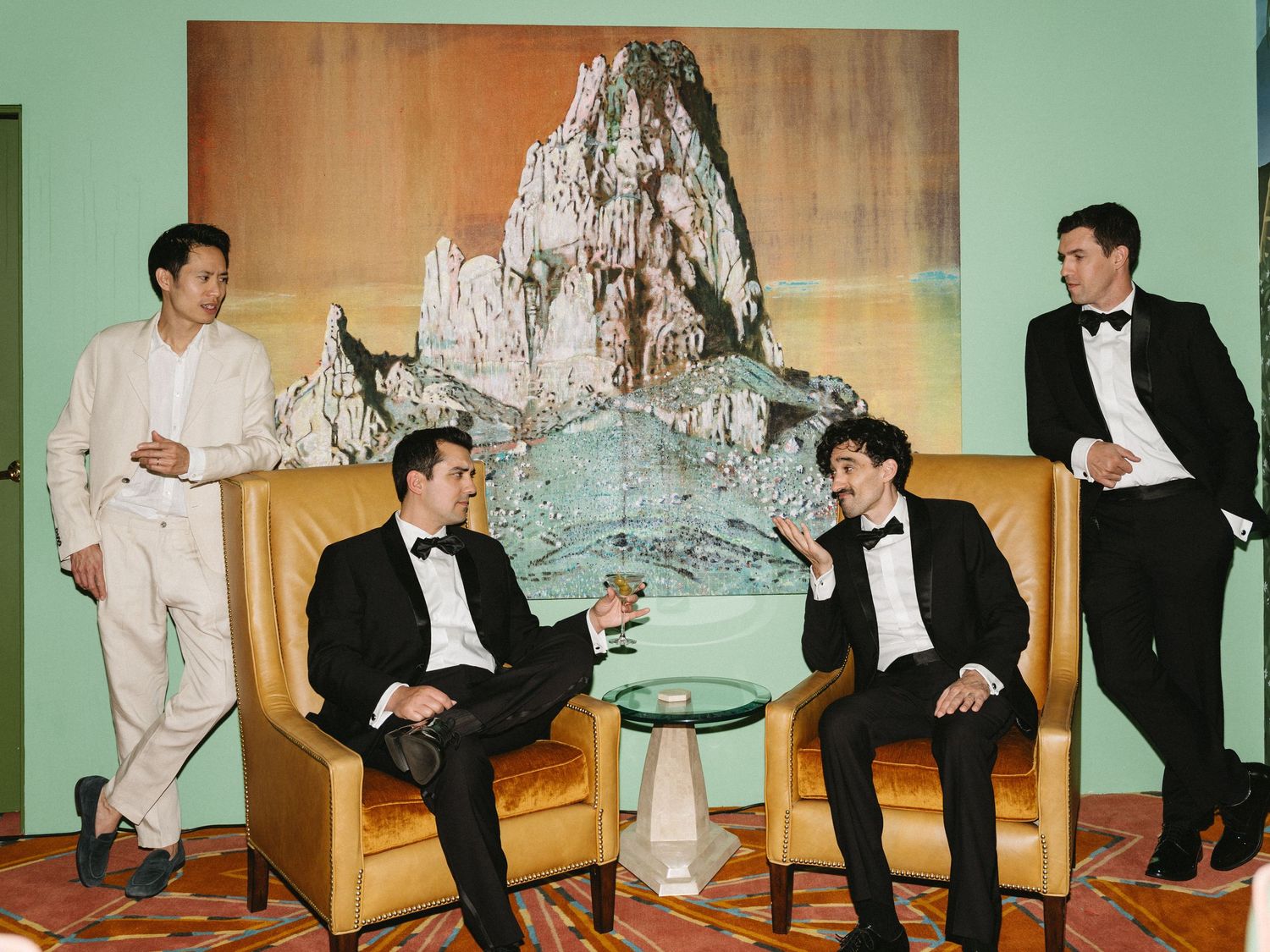 Four men in formal attire pose in an elegant room with a mountain painting backdrop and gold chairs.