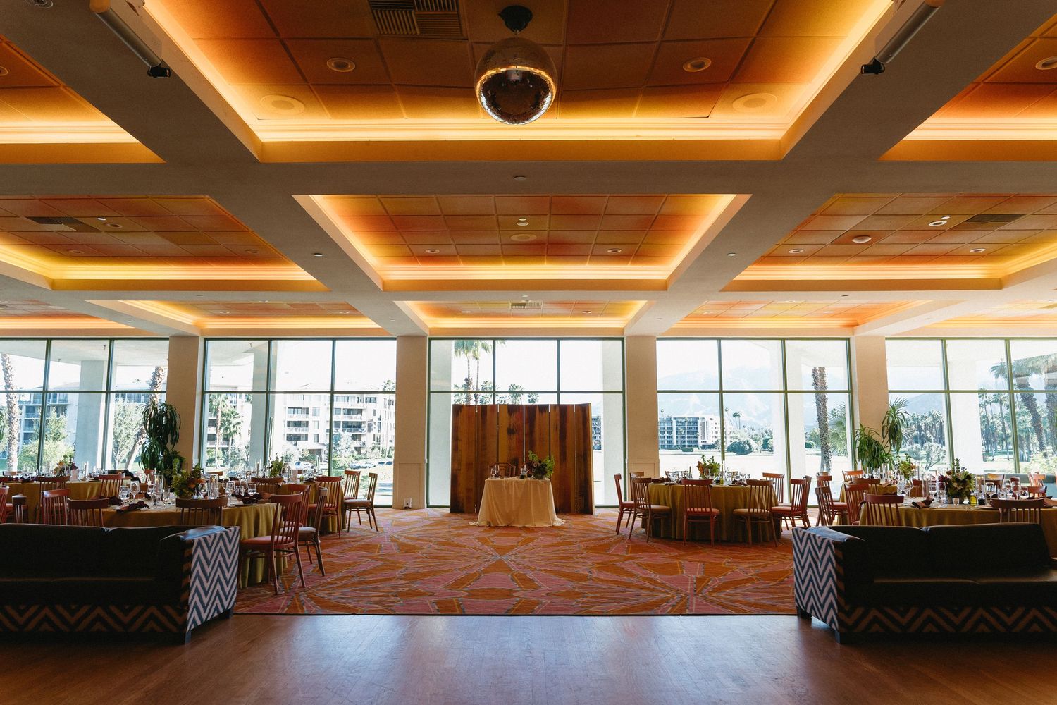 Elegant banquet hall with coffered ceiling, warm lighting, round tables, and large windows overlooking outdoor scenery.