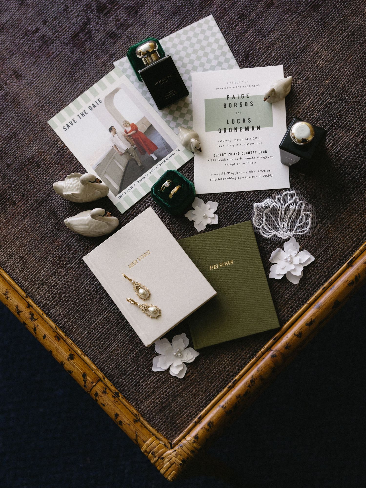 Elegant wedding flat lay with green invitations, pearl earrings, rings, and floral details on wooden surface.