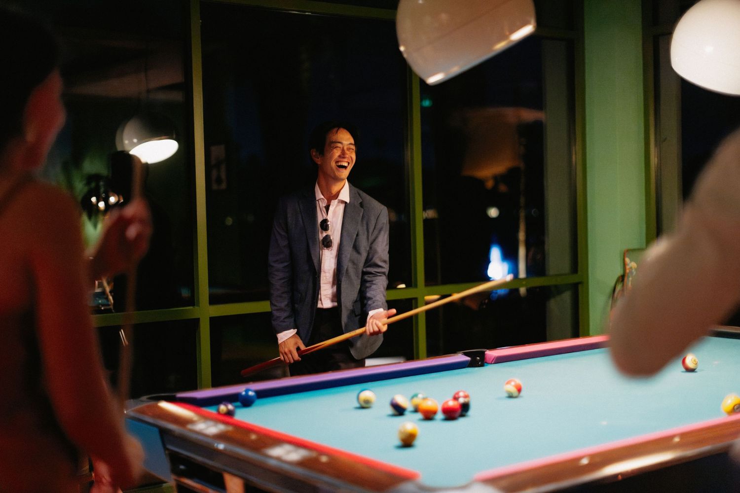 Man laughing while playing pool billiards in a stylish bar, holding a cue stick near a colorful billiard table.