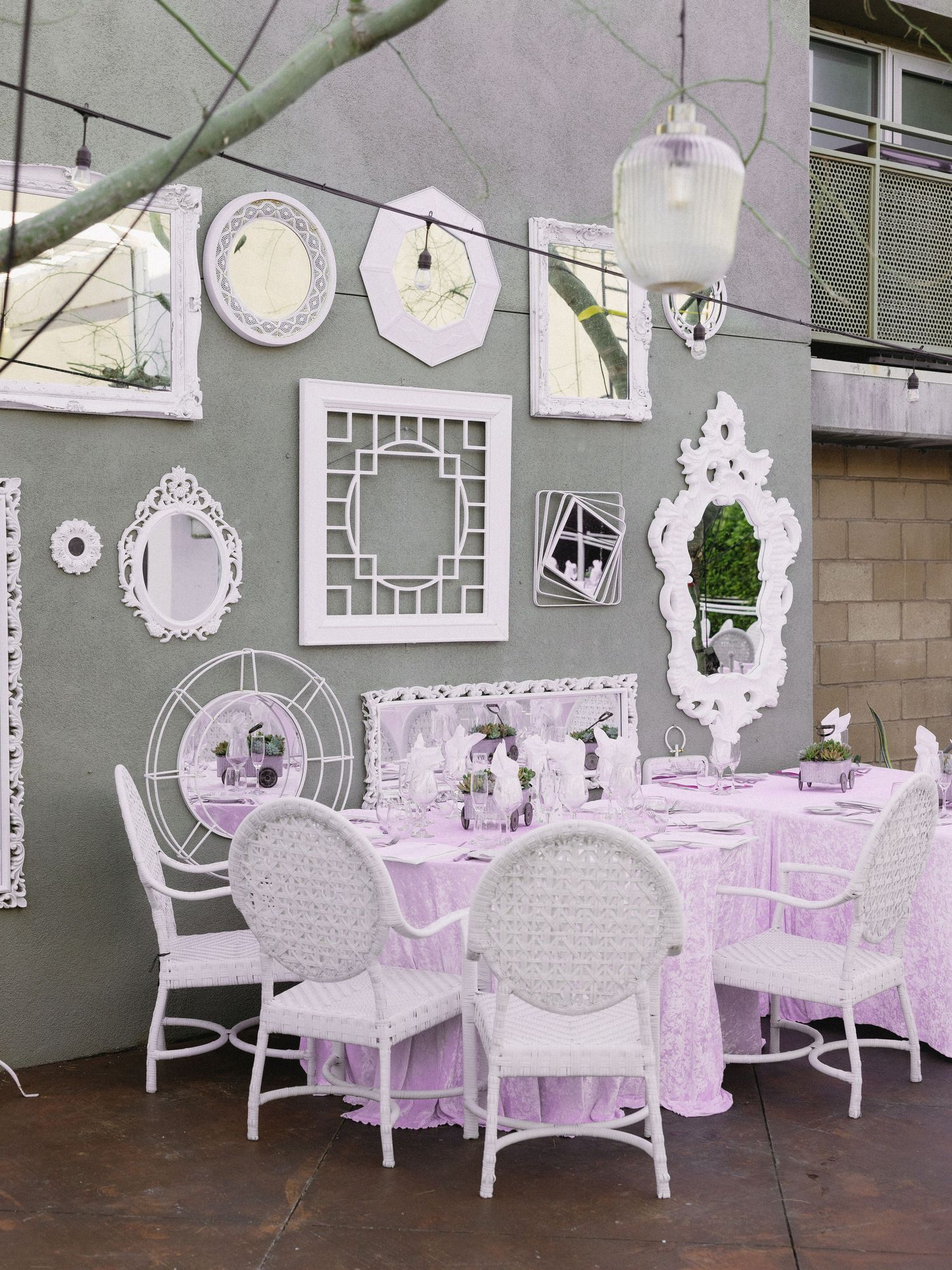Elegant outdoor dining setup with white wicker chairs, pink tablecloths, and ornate white mirrors on a gray wall.