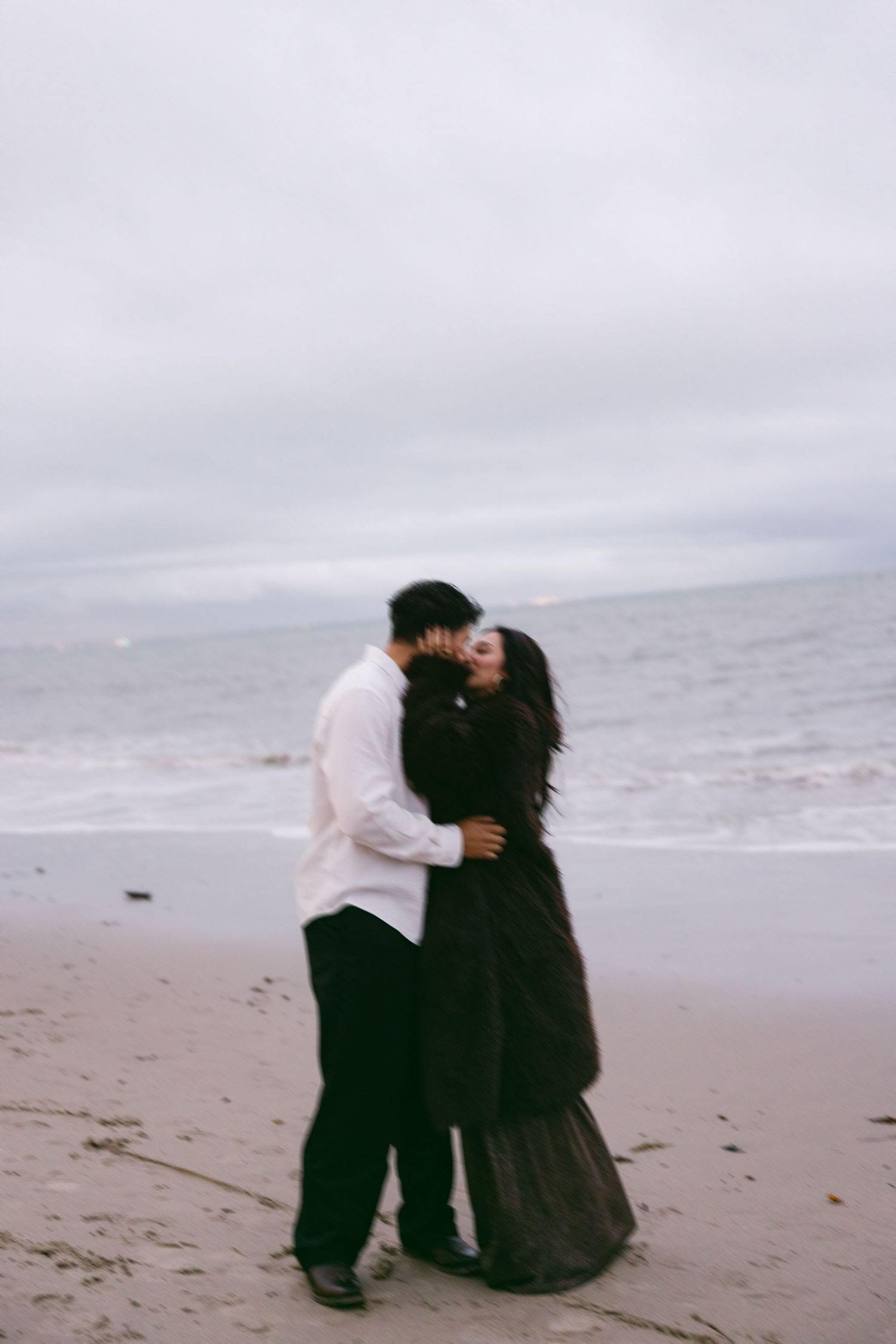 Couple embracing on cloudy beach with waves in background.
