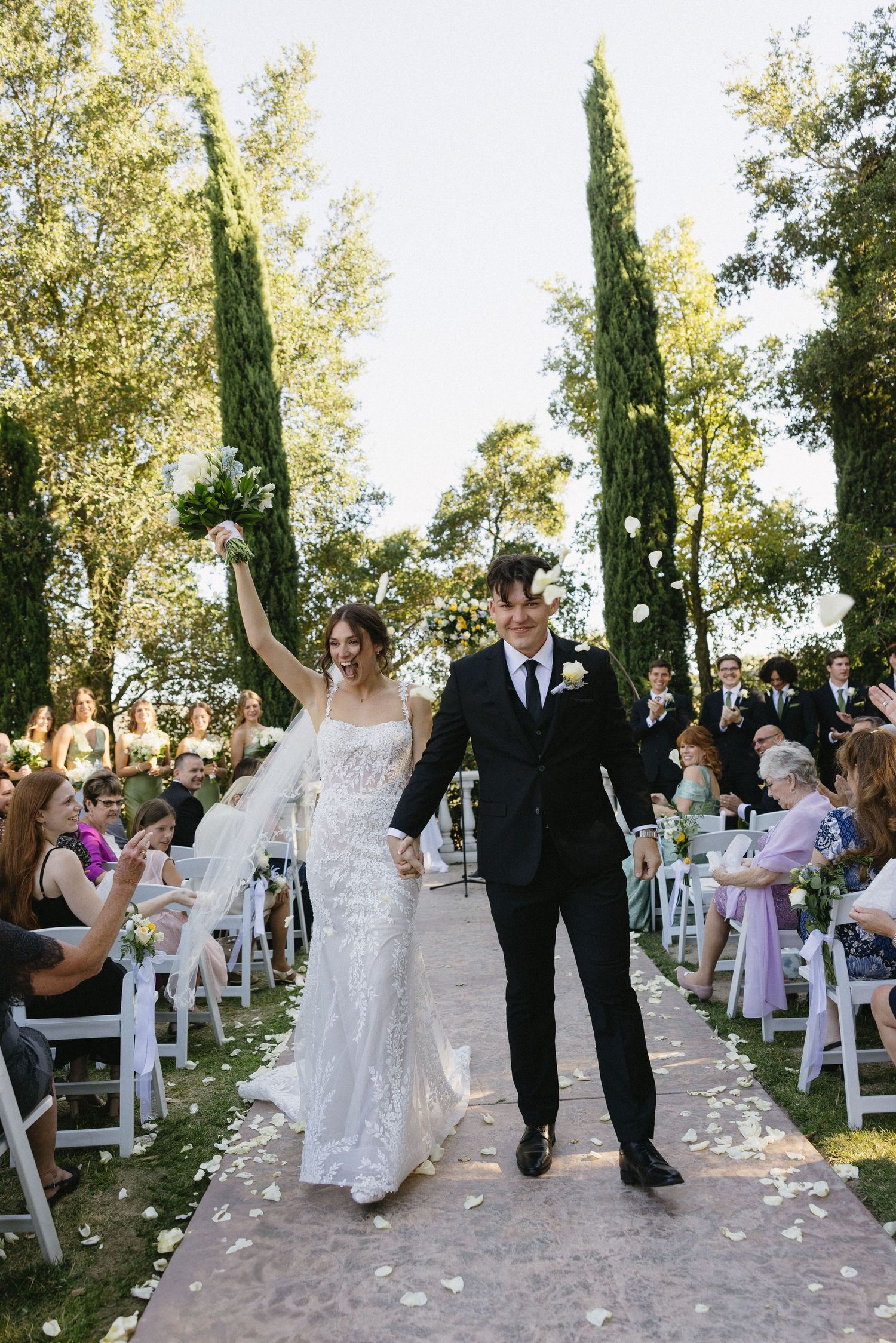 Joyful bride and groom walk down outdoor aisle after wedding ceremony, surrounded by cypress trees and cheering guests.