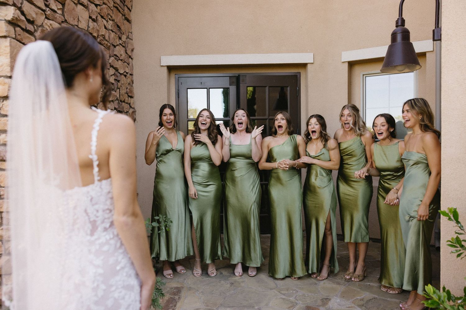 Bridesmaids in sage green satin dresses react with joy and excitement as bride in white lace gown reveals her look.