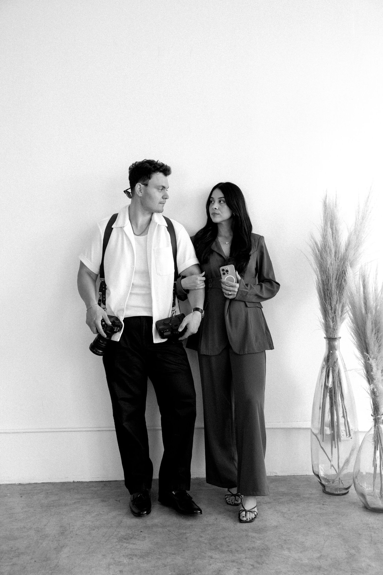 Black and white photo of a young man and woman posing against a white wall, man holding camera, woman holding phone.