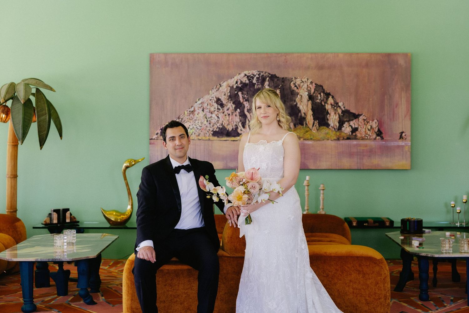 Bride and groom pose on velvet sofa before green wall with mountain painting at stylish wedding venue.