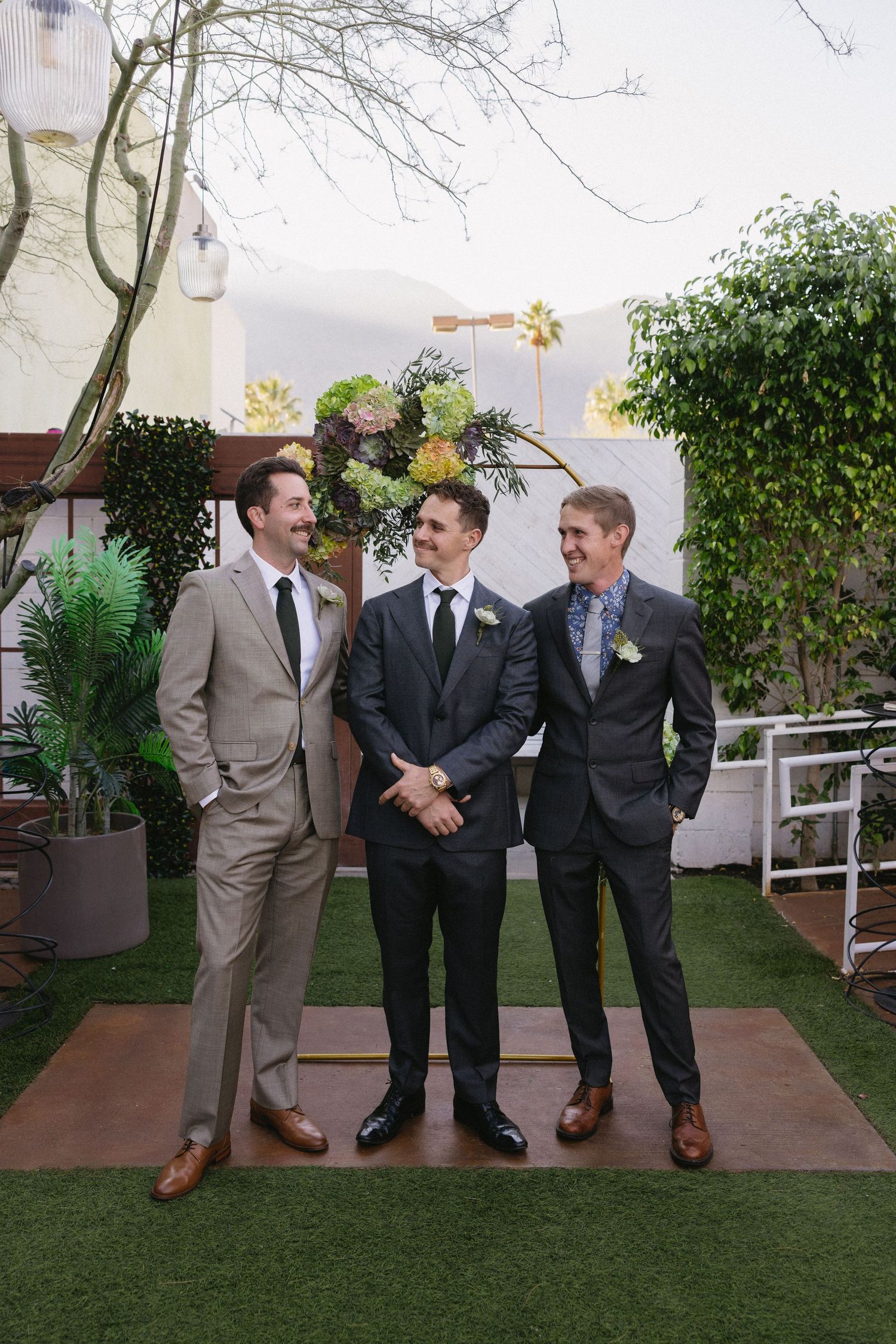 Groom and two groomsmen in suits smiling together in front of floral arch at outdoor wedding ceremony.