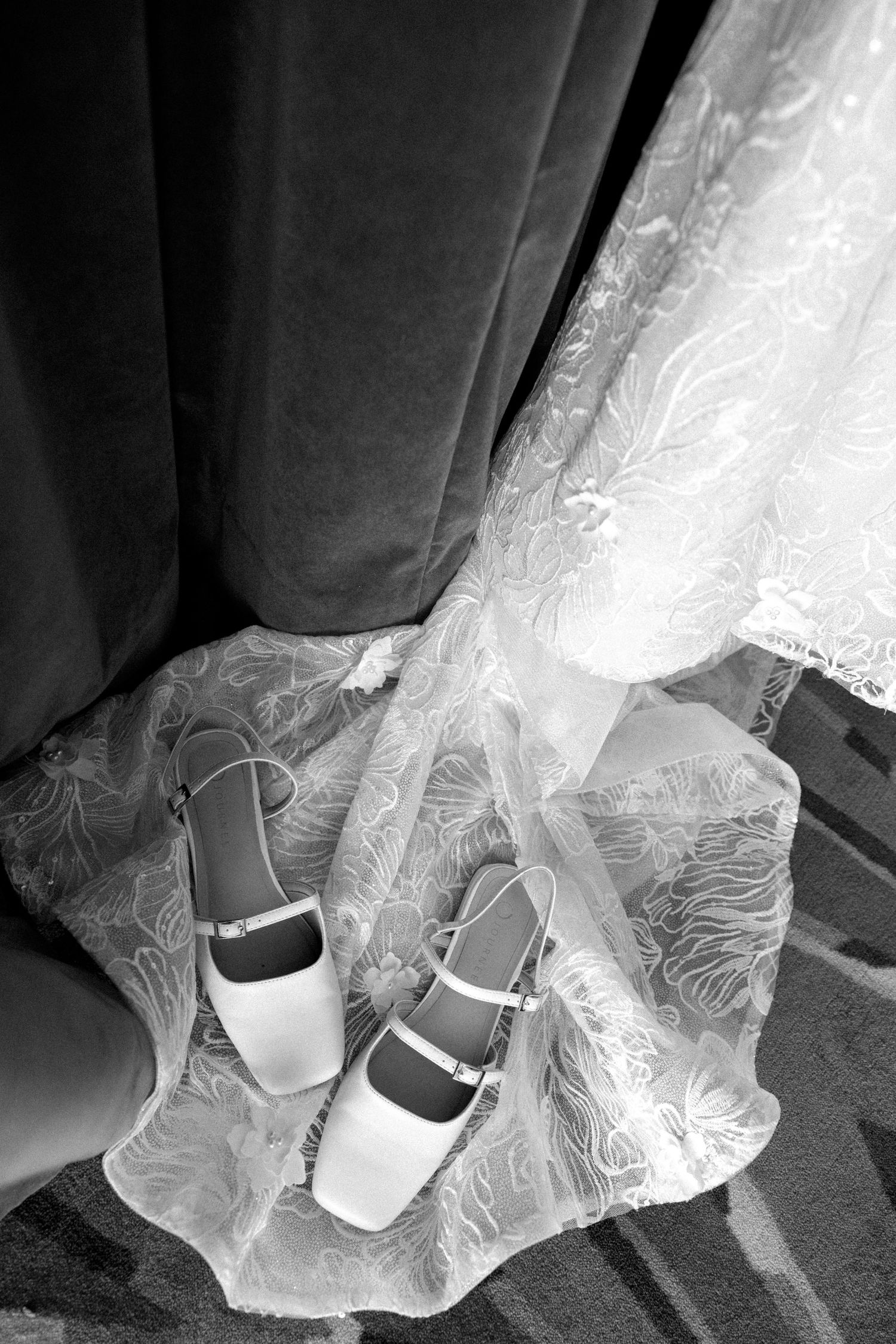 Black and white photo of elegant bridal shoes resting on lace wedding dress fabric near a curtain.