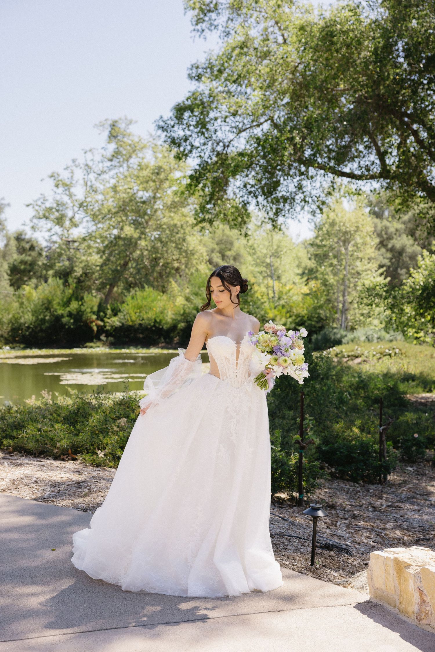 Bride in a strapless ball gown with puff sleeves holds a floral bouquet outdoors near a serene pond.