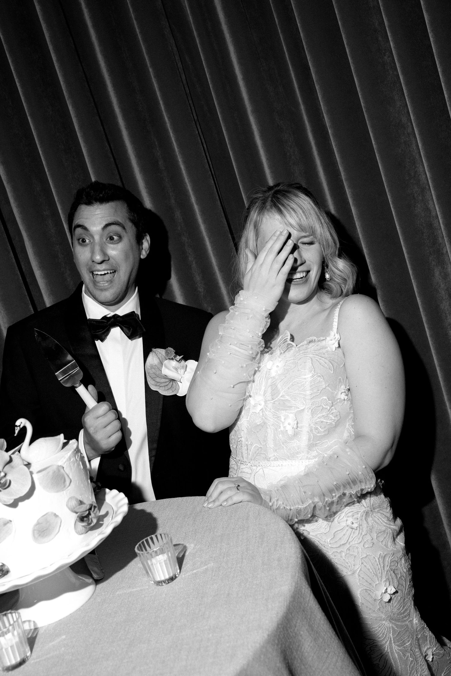 Black and white photo of a laughing bride and groom cutting their wedding cake at a reception.