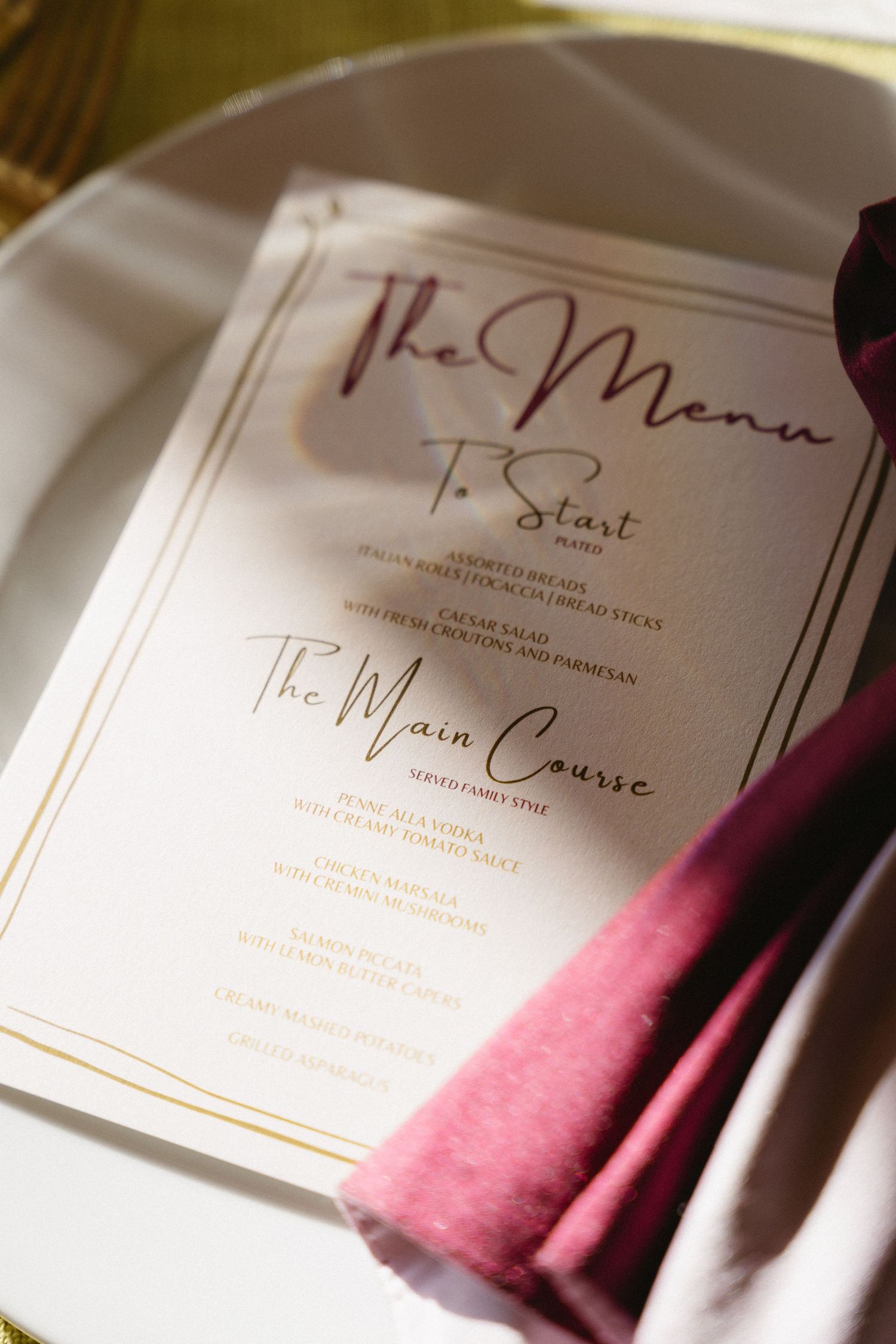 Elegant wedding menu card in gold and burgundy script on white plate with pink linen napkin.