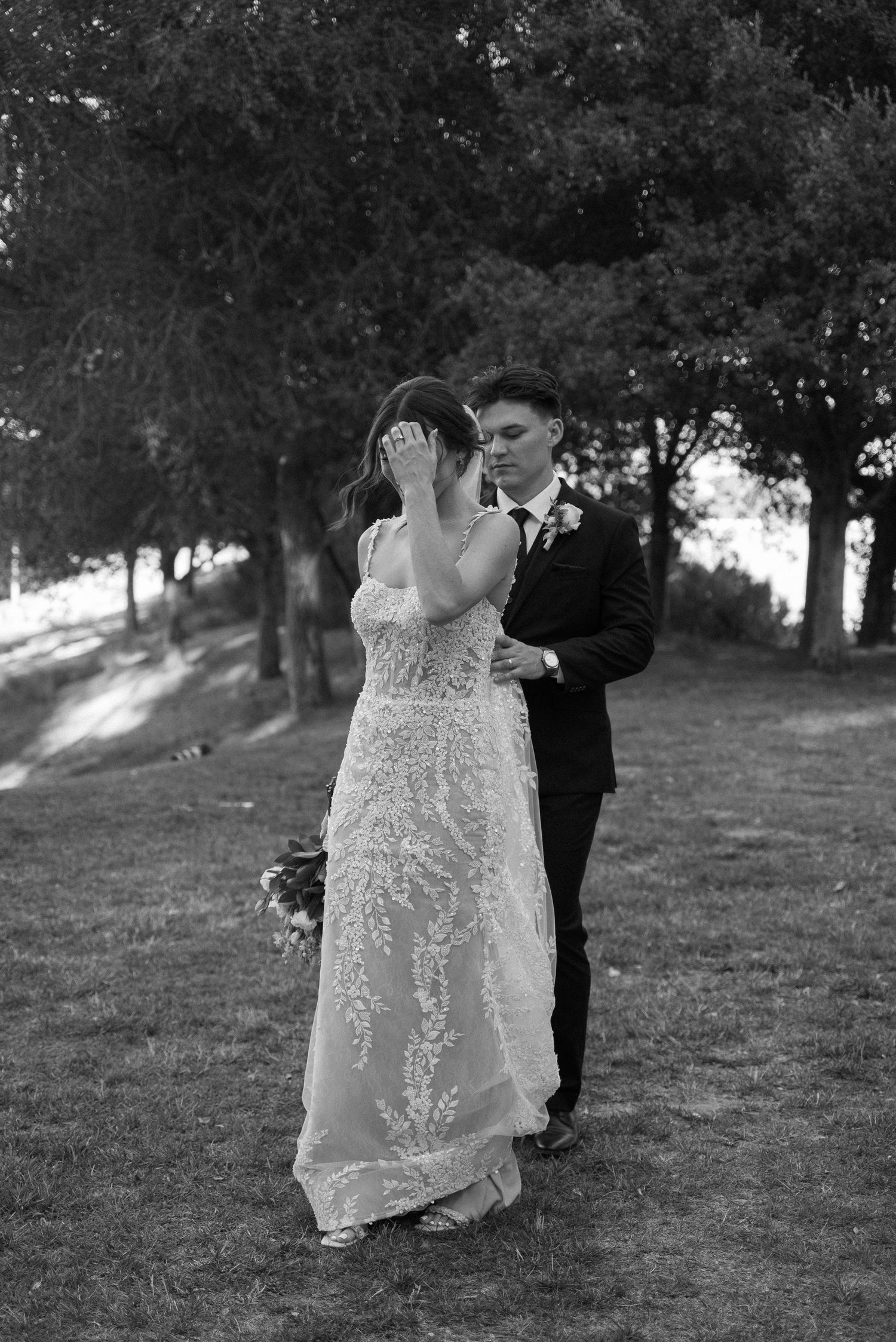 Romantic couple poses in flowing lace wedding dress among trees at sunset.