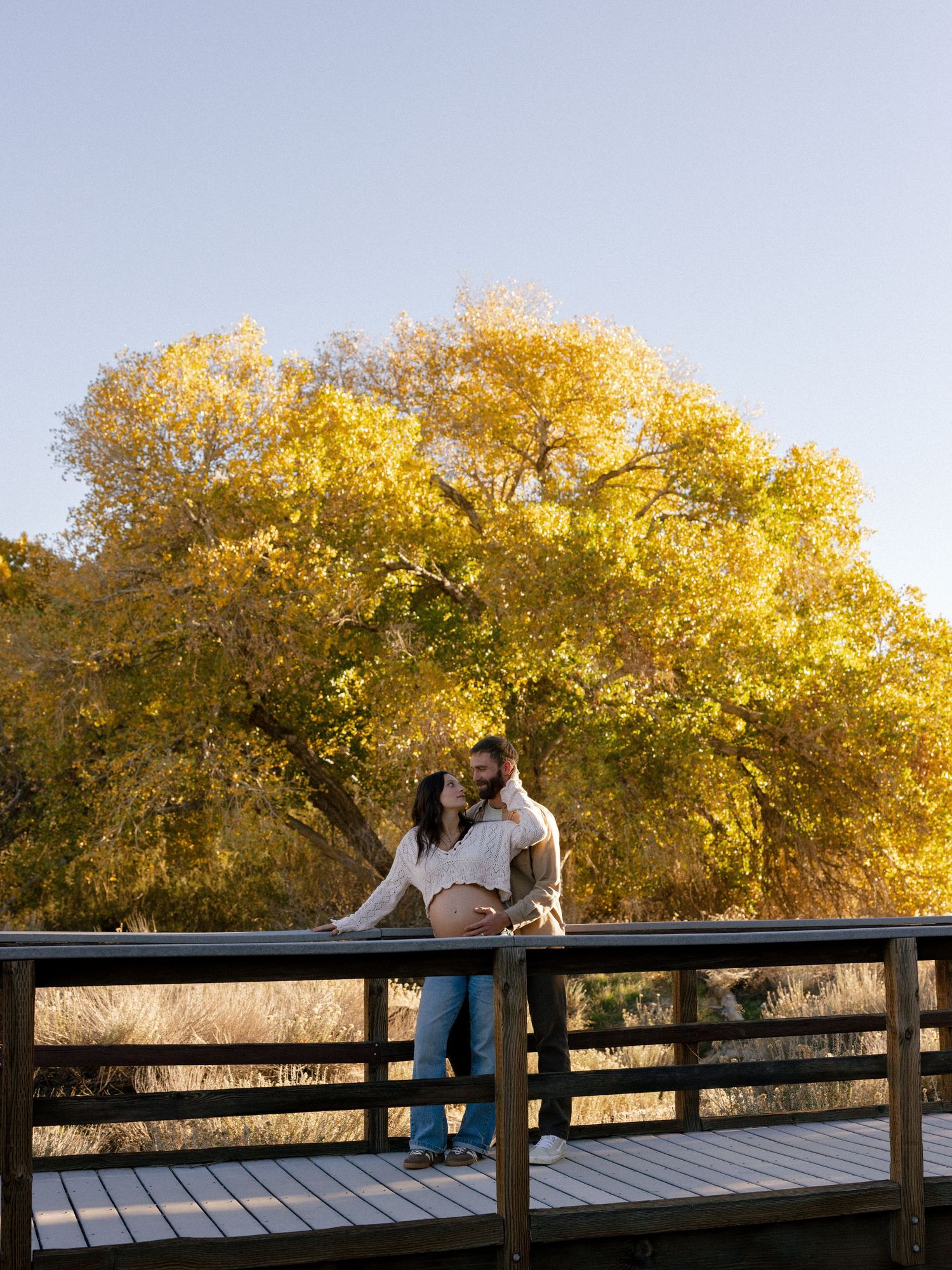 Couple shares tender moment on wooden bridge with golden autumn trees in background.