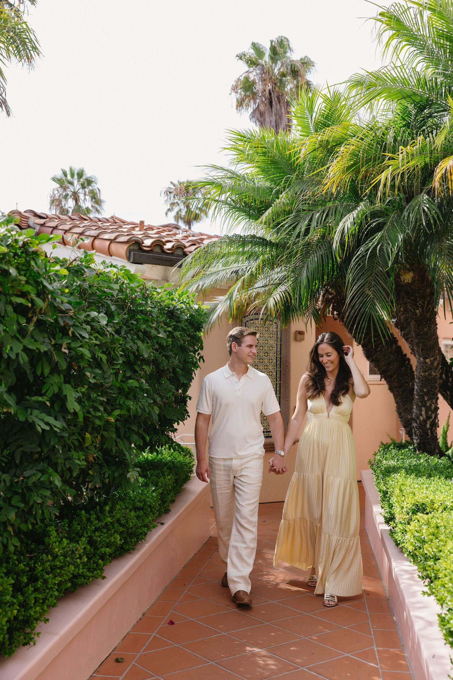 Couple holding hands walking along a tropical resort pathway lined with palm trees and lush greenery.