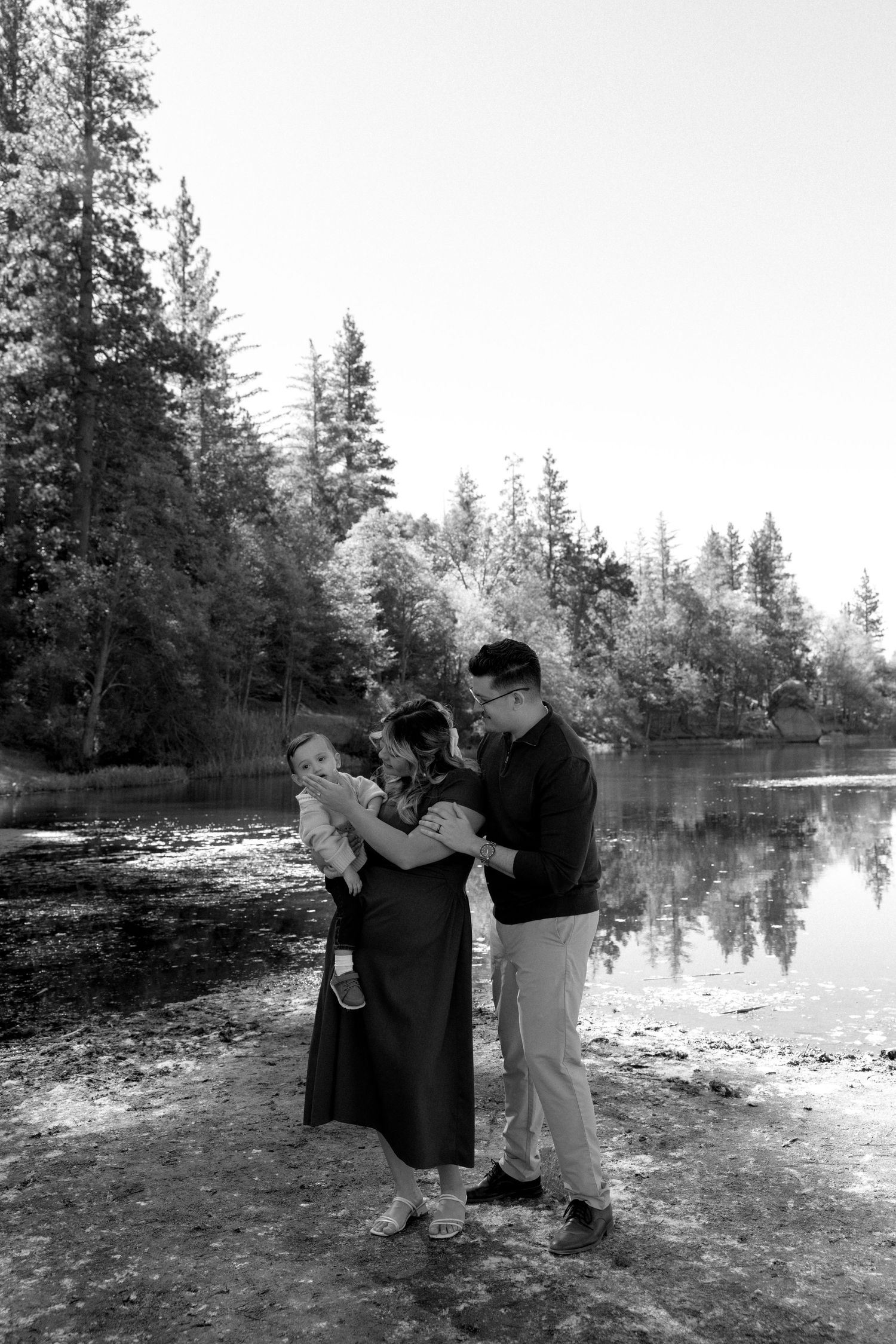 Black and white photo of a family of four standing by a reflective lake surrounded by tall pine trees.