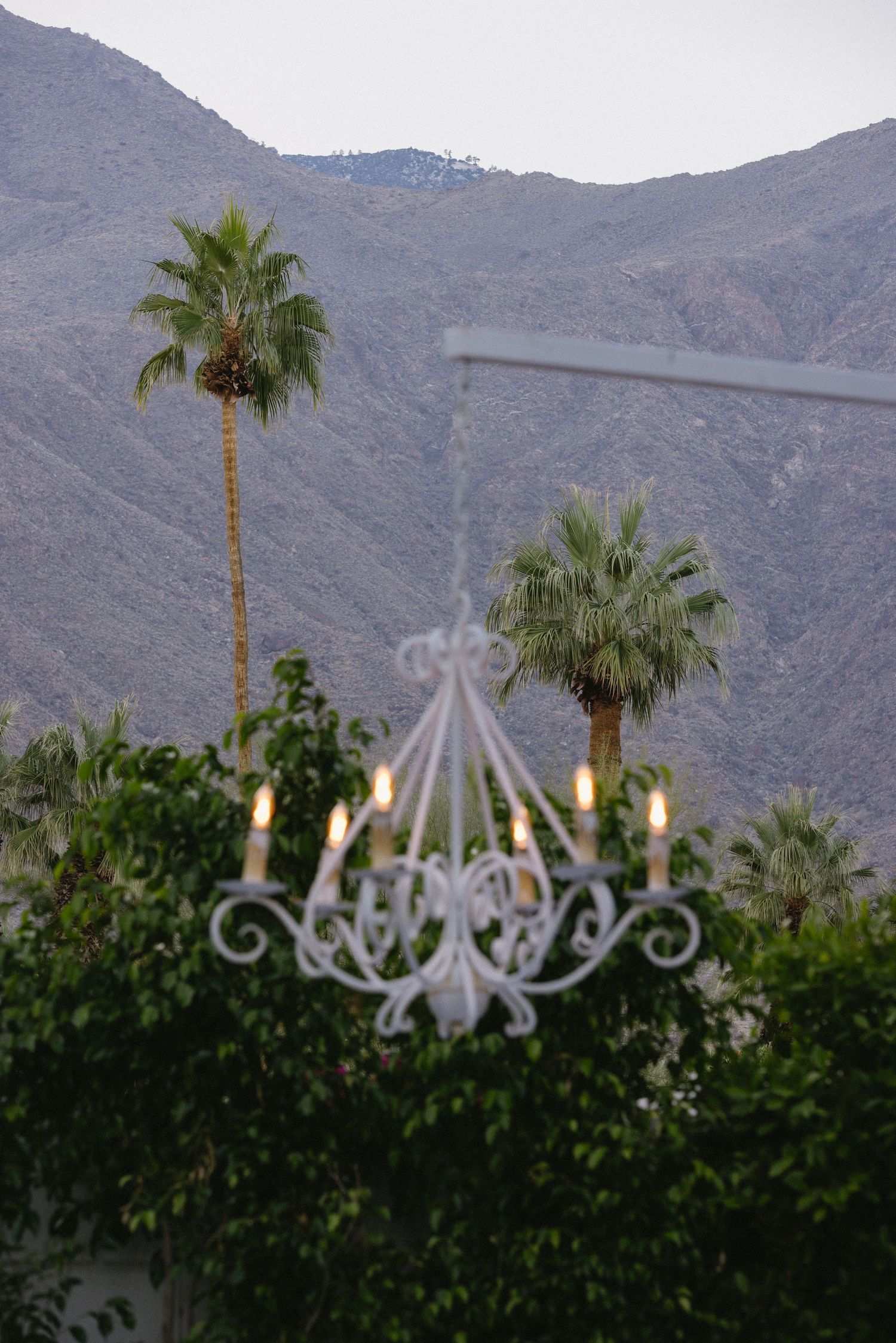 White wrought iron chandelier with lit candles outdoors, framed by palm trees and mountain backdrop at dusk.