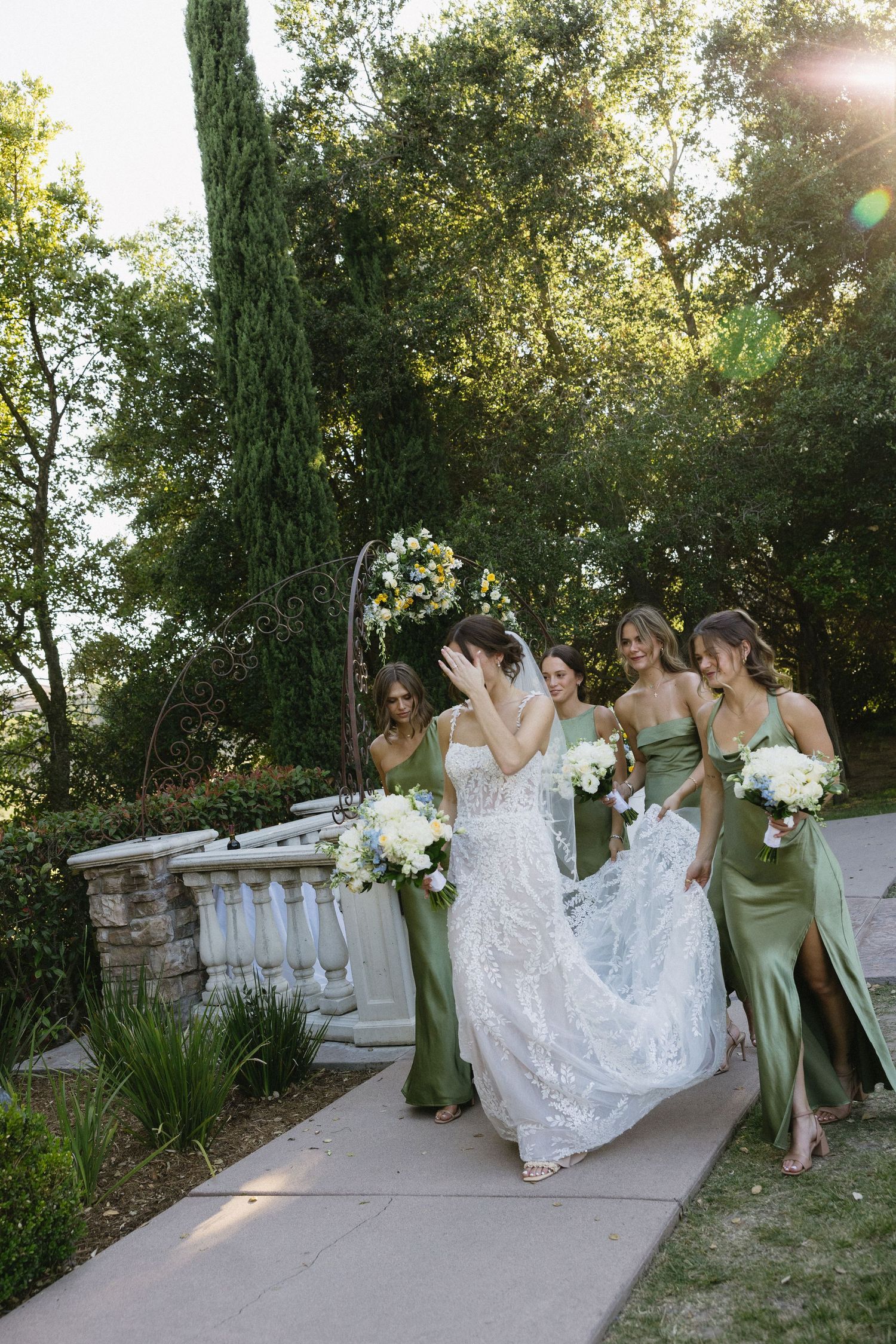Bride in lace gown walks with bridesmaids in sage green dresses carrying white bouquets in a garden.
