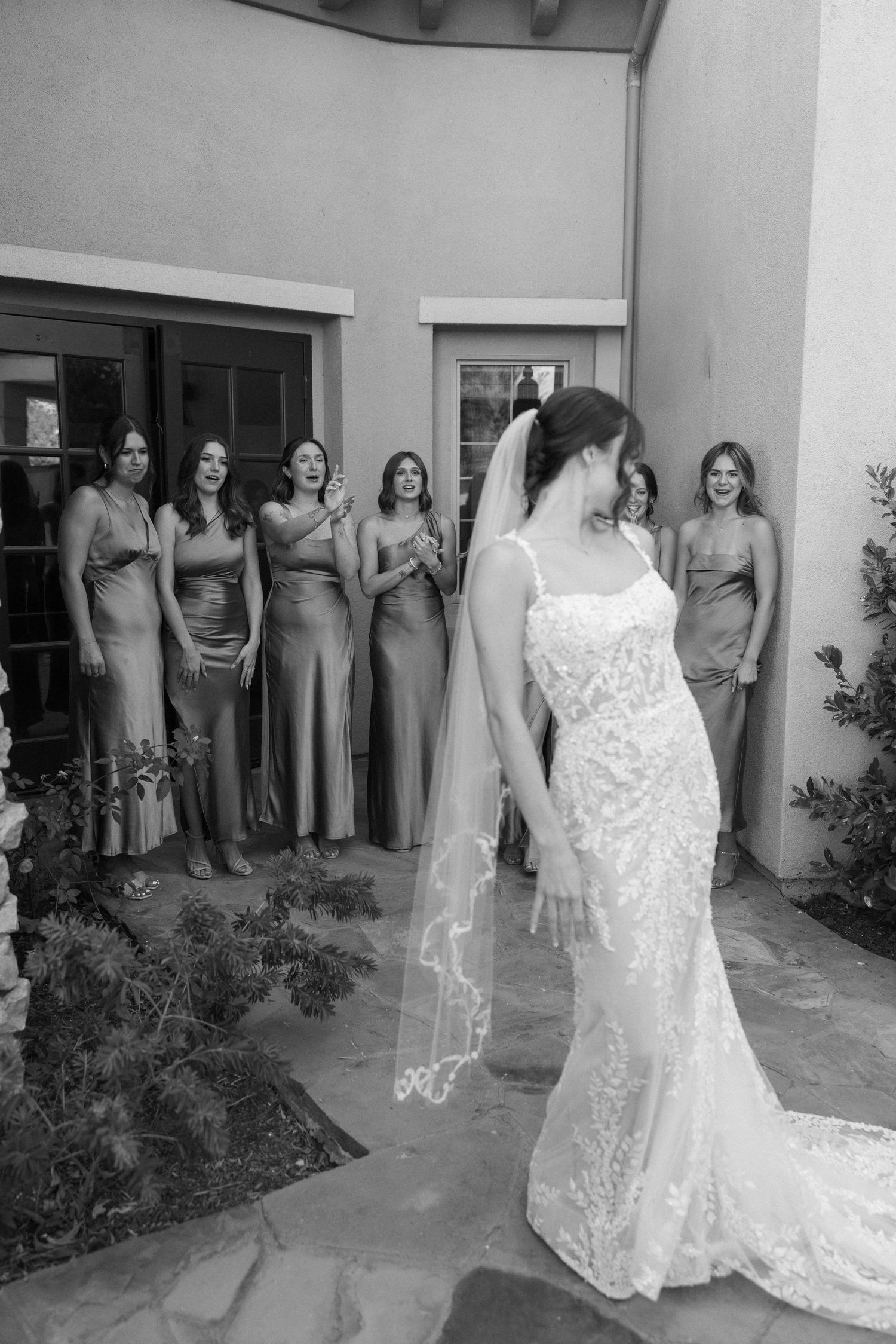 Bride in lace gown and veil reveals herself to bridesmaids in satin dresses during outdoor first look.