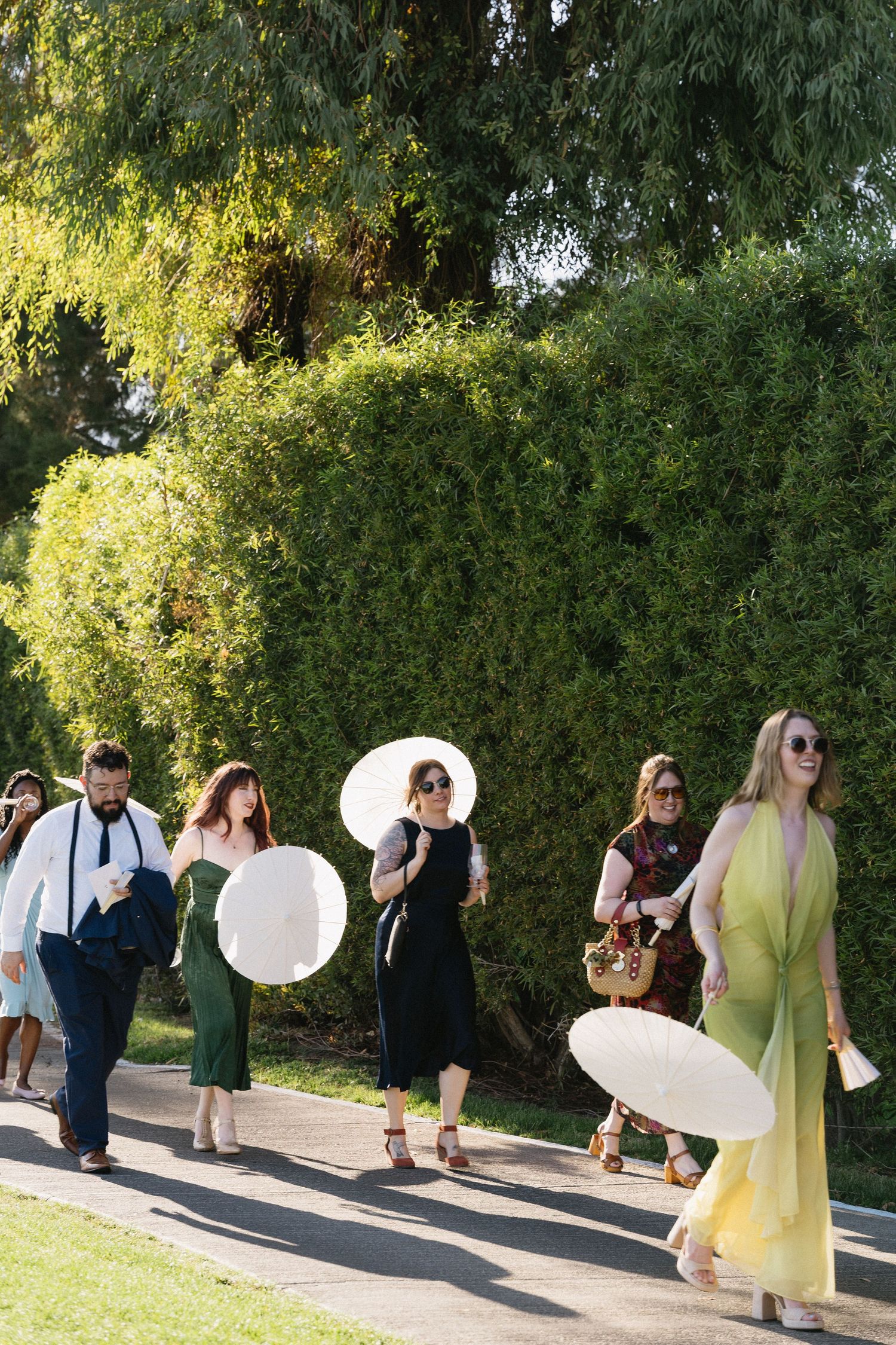 Elegantly dressed wedding guests walk along a garden path holding white parasols in sunny outdoor setting.
