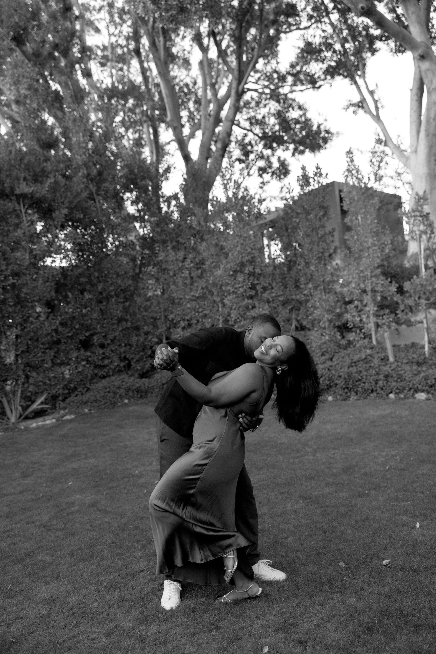 Palm Springs Black and white photo of a couple dancing and laughing together on a green lawn with trees in the background.