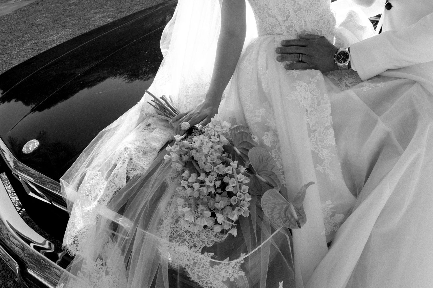Black and white photo of a bride in lace gown holding a floral bouquet beside a car on her wedding day.
