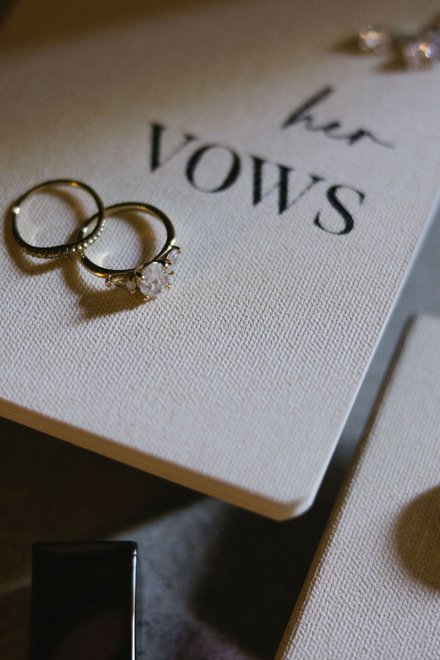 Gold wedding rings and diamond engagement ring resting on a white vow book labeled her VOWS.