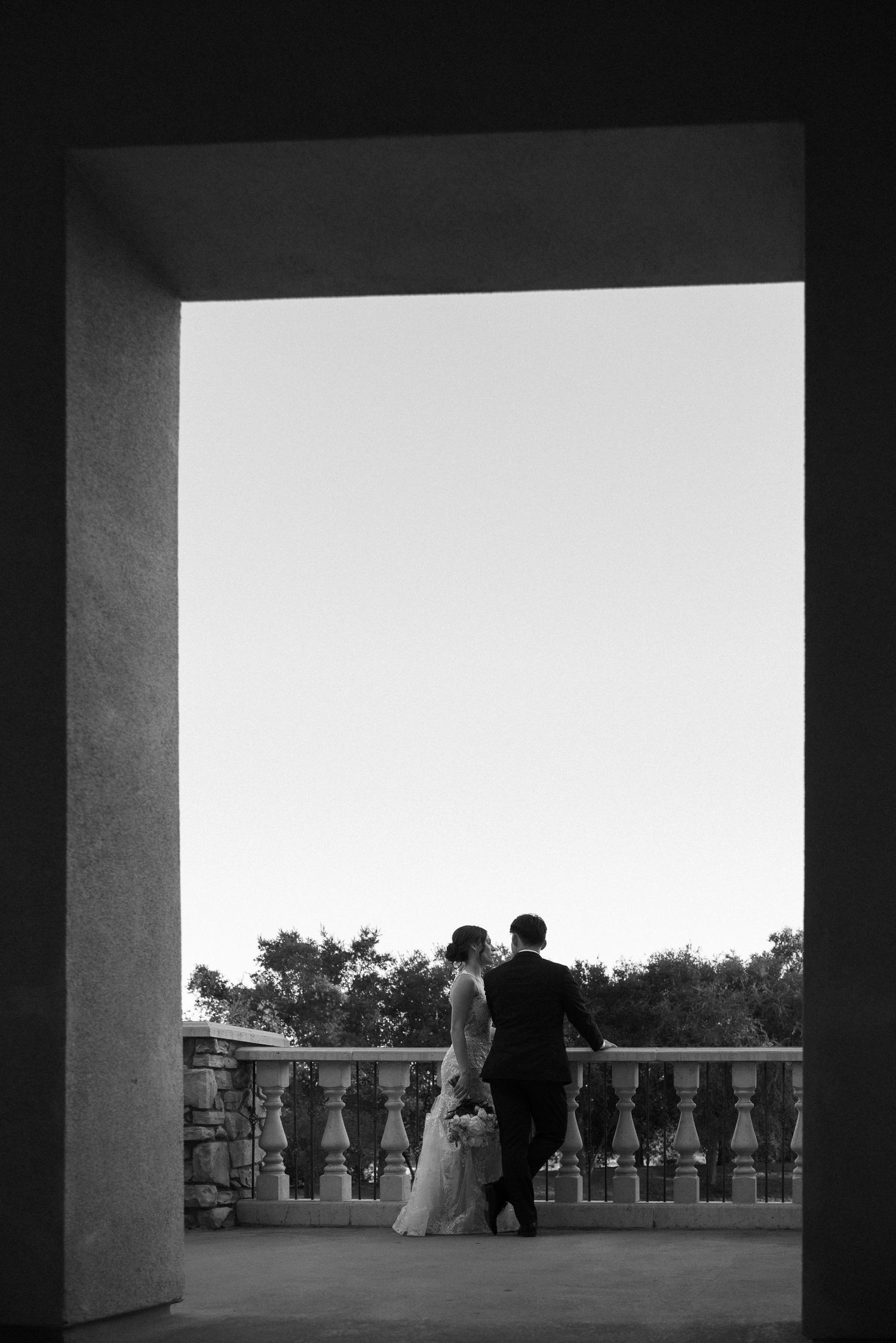 Black and white photo of a bride and groom standing on an elegant balcony framed by a stone archway.