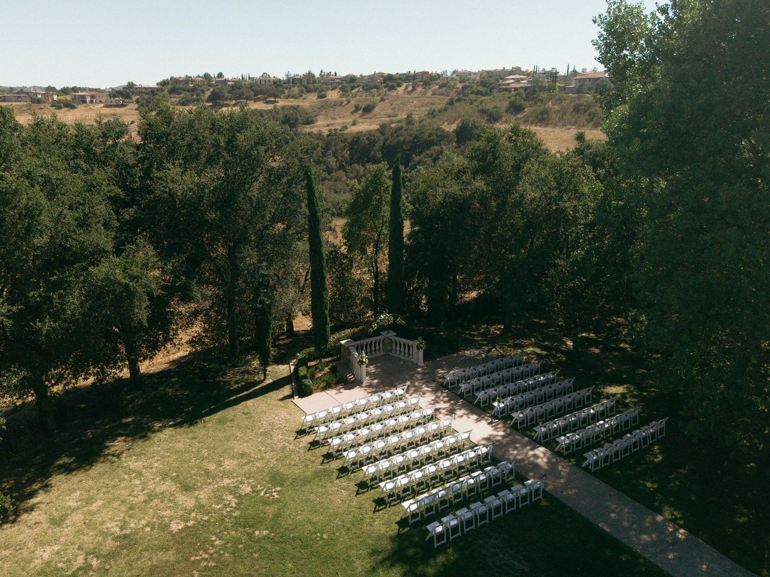 Aerial view of an elegant outdoor wedding ceremony setup with white chairs on a green lawn surrounded by trees.