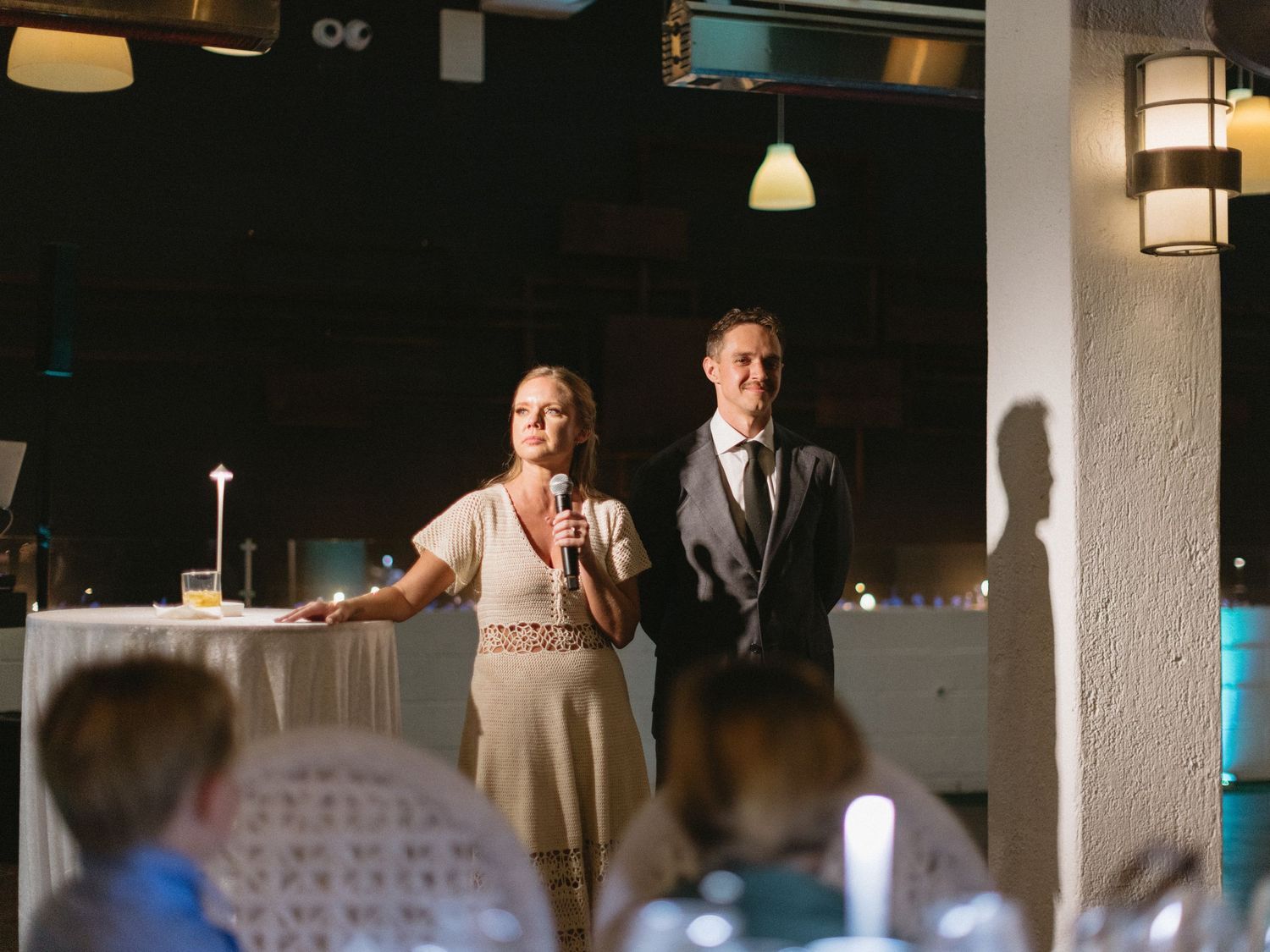 A woman speaks at a wedding reception podium while a man in a suit stands beside her in a dimly lit venue.