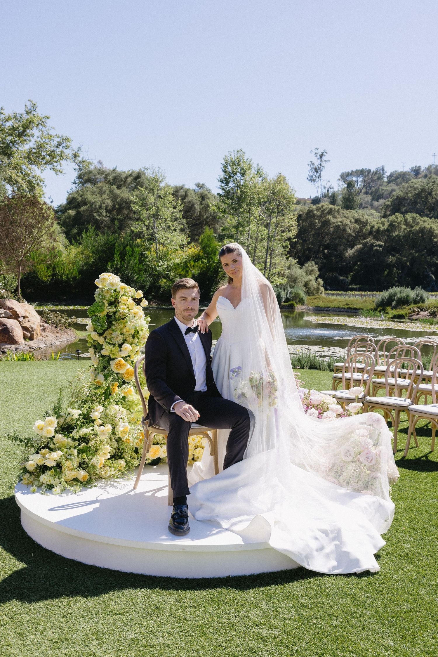 Bride and groom pose on a circular platform surrounded by yellow floral arrangements at outdoor wedding.