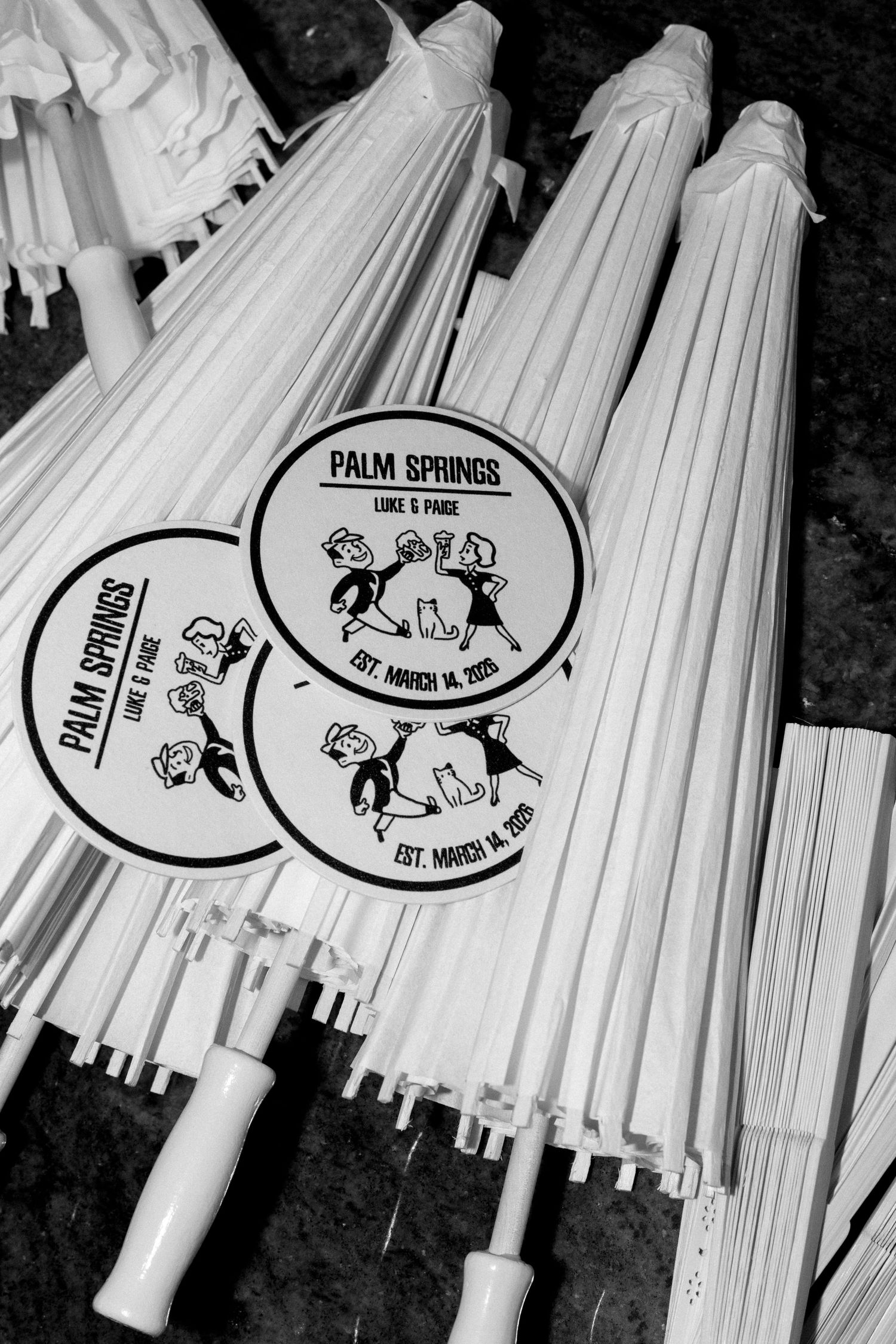 Black and white photo of white parasols with Palm Springs Luke and Paige Est. March 14, 2025 wedding logos.