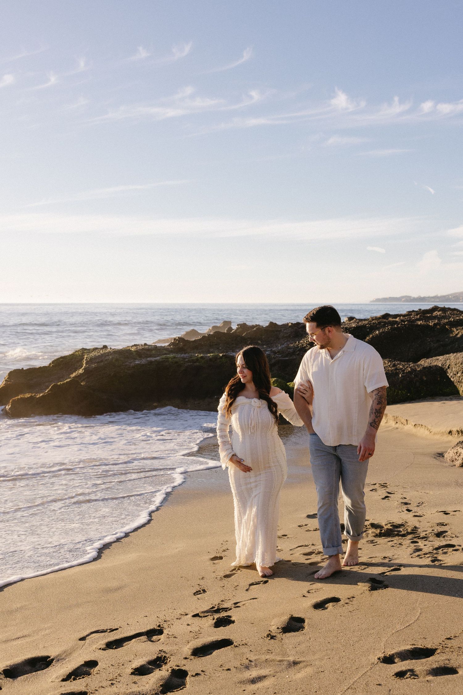 A couple walks together on a scenic beach at sunset with rocky cliffs in the background.