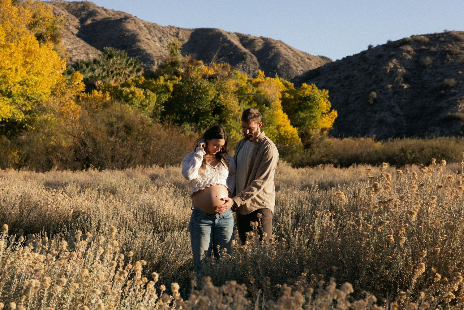 Two people walk through a desert field with mountains and autumn trees in the background.