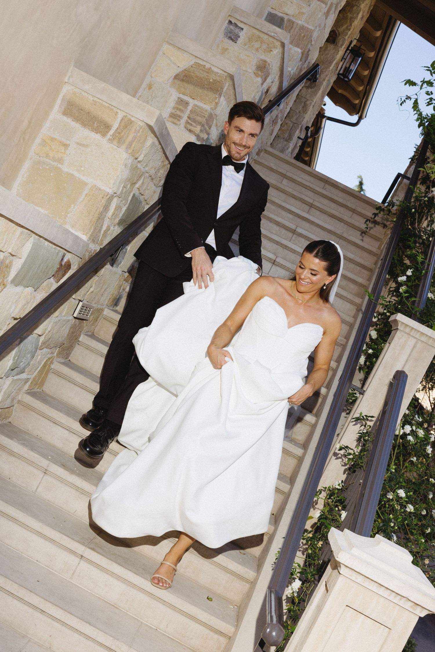 Bride in strapless white gown and groom in black tuxedo descending stone staircase at elegant wedding venue.