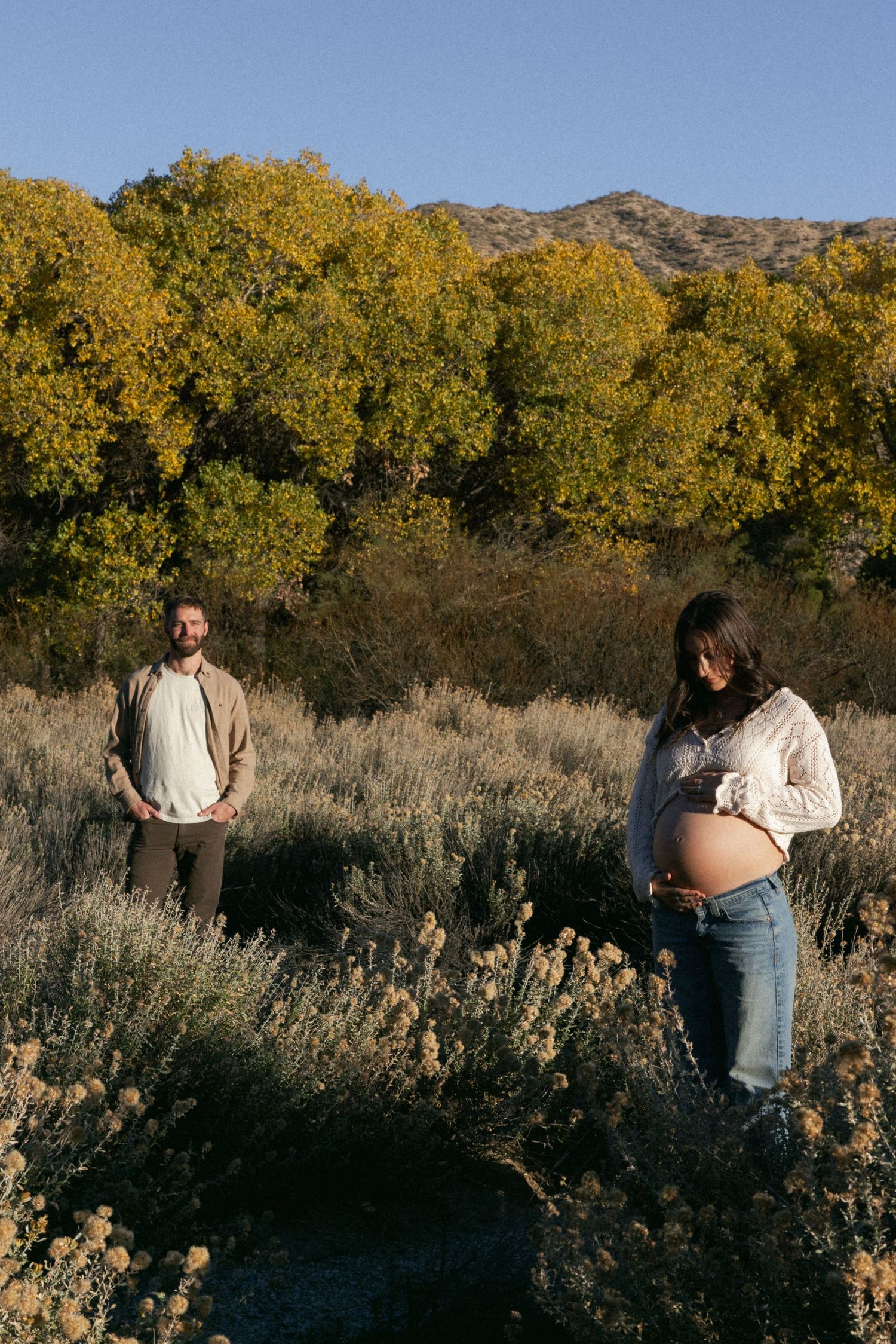 A couple stands apart in a desert field with golden trees and mountains in the background.