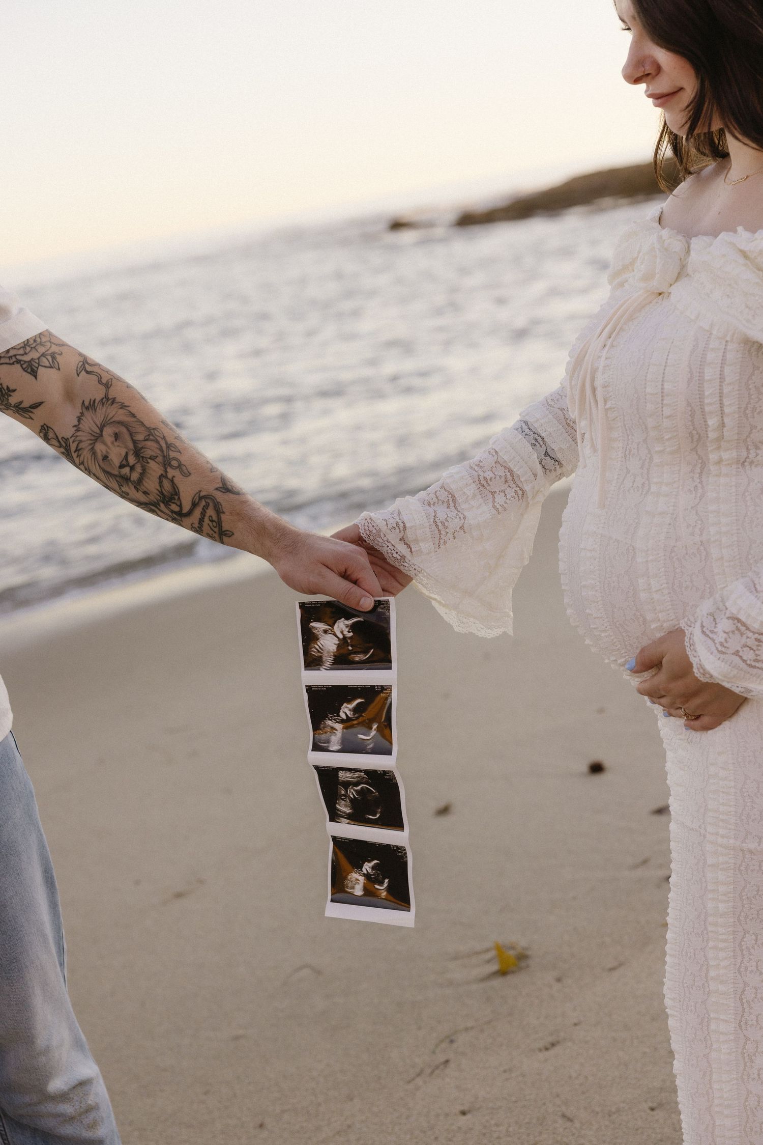 Hands holding ultrasound photos on a sunny beach with ocean waves in the background.