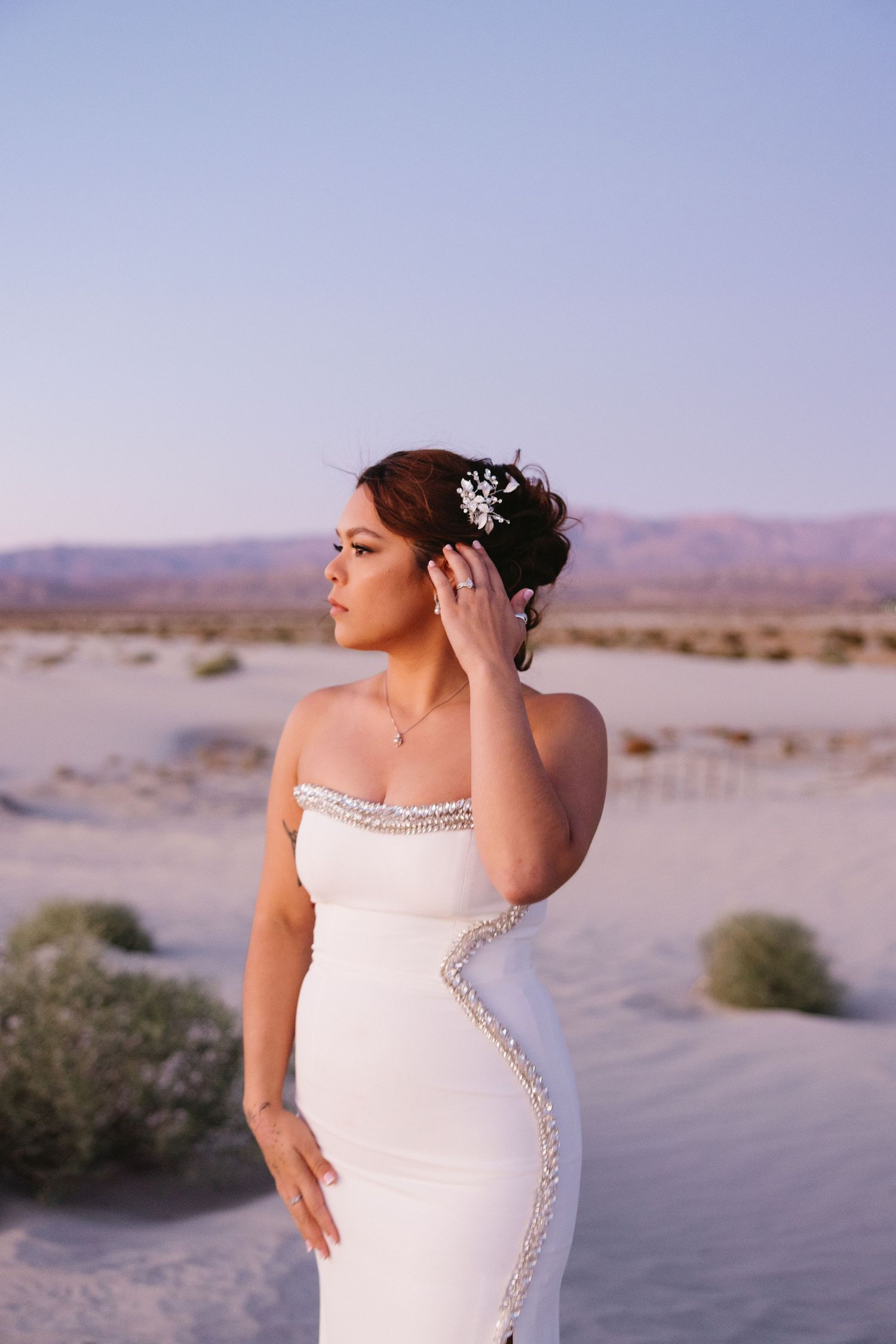 Bride in fitted white dress with crystal details poses against desert landscape at sunset.