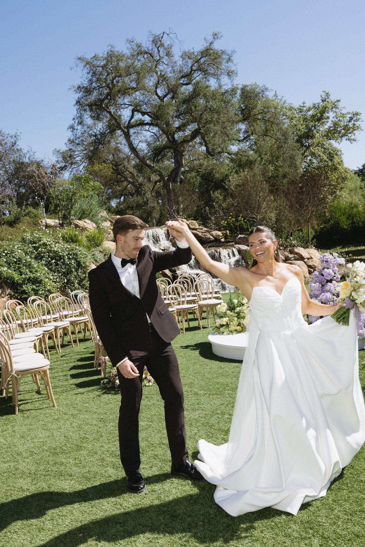Bride and groom celebrate joyfully on green lawn with waterfall and wedding chairs in background.